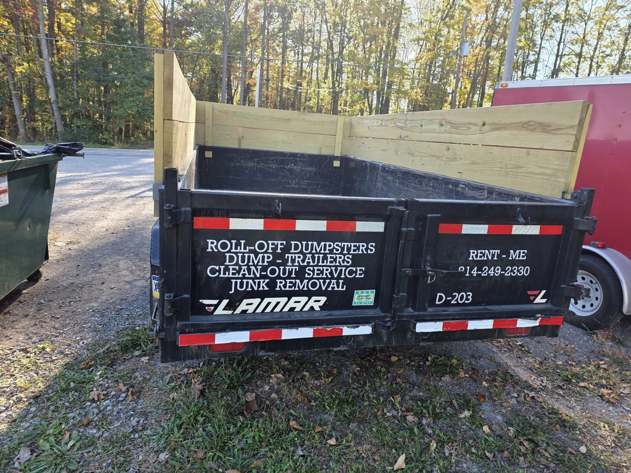 Black trailer with red and white reflective stripes around the bottom, parked on a grassy area beside a road during fall, with trees and sunlight in the background.