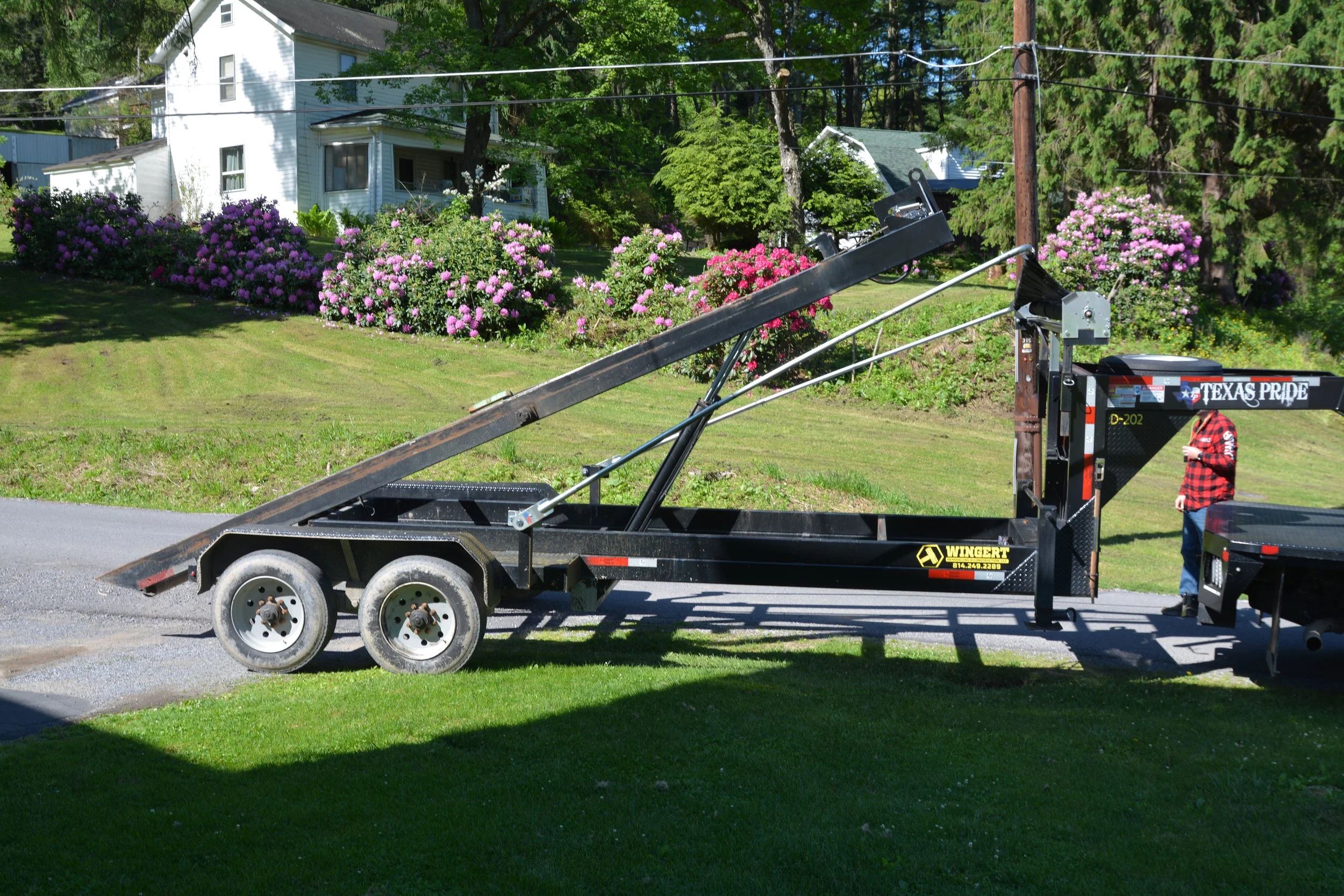 A flatbed trailer parked on a grassy lawn with a mechanical lift and a person in a red and black plaid shirt standing nearby. In the background, there are blooming pink and purple flowers, a white house, trees, and utility poles.