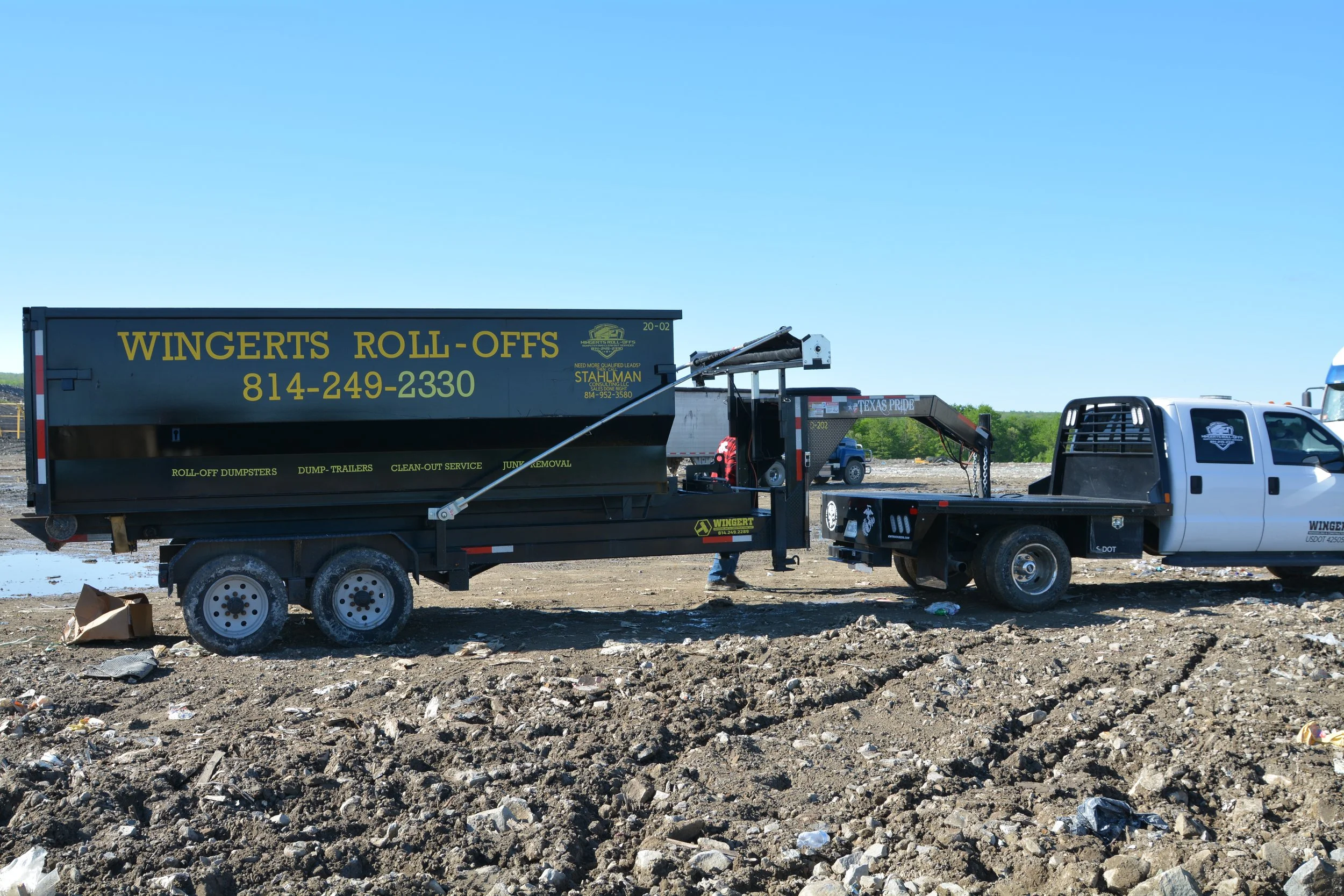 A construction site with a truck and trailer labeled Wingerts Roll-Offs, with a clear blue sky overhead.