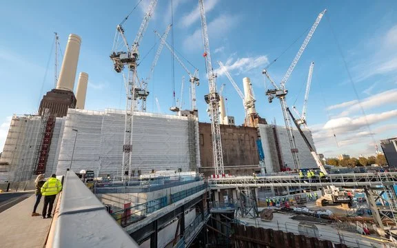 Construction site with workers and multiple cranes building a large structure under a blue sky.