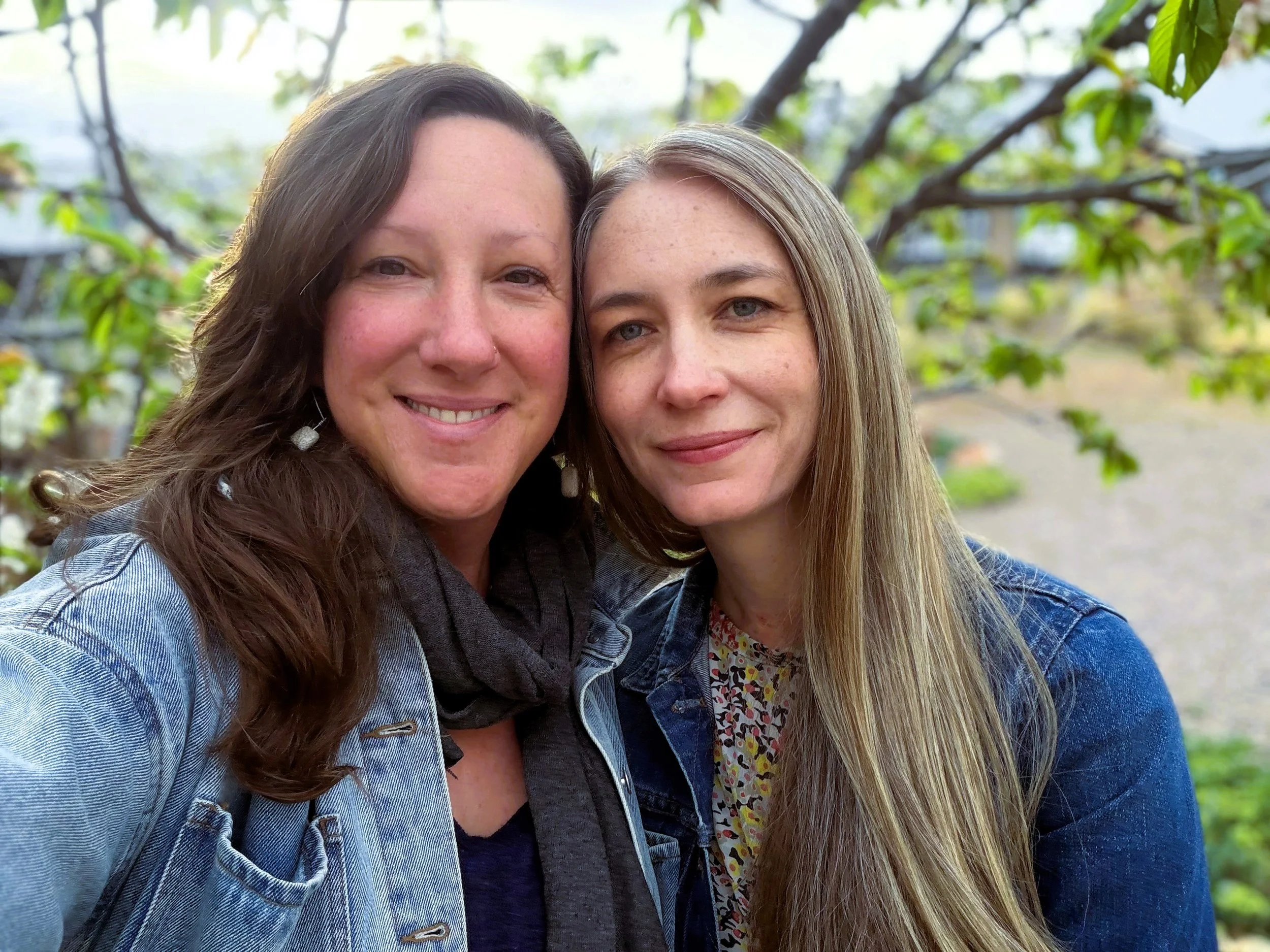 Two women smiling close together outdoors in front of a tree with green leaves.