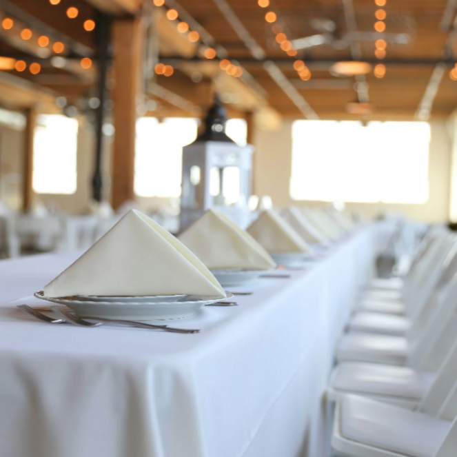 A long banquet table set with white tablecloths, folded white napkins, and silverware in a decorated banquet hall with string lights and windows.