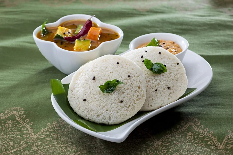 Idlis served on a white plate with green leaves and black pepper, accompanied by sambar and chutney in separate bowls.