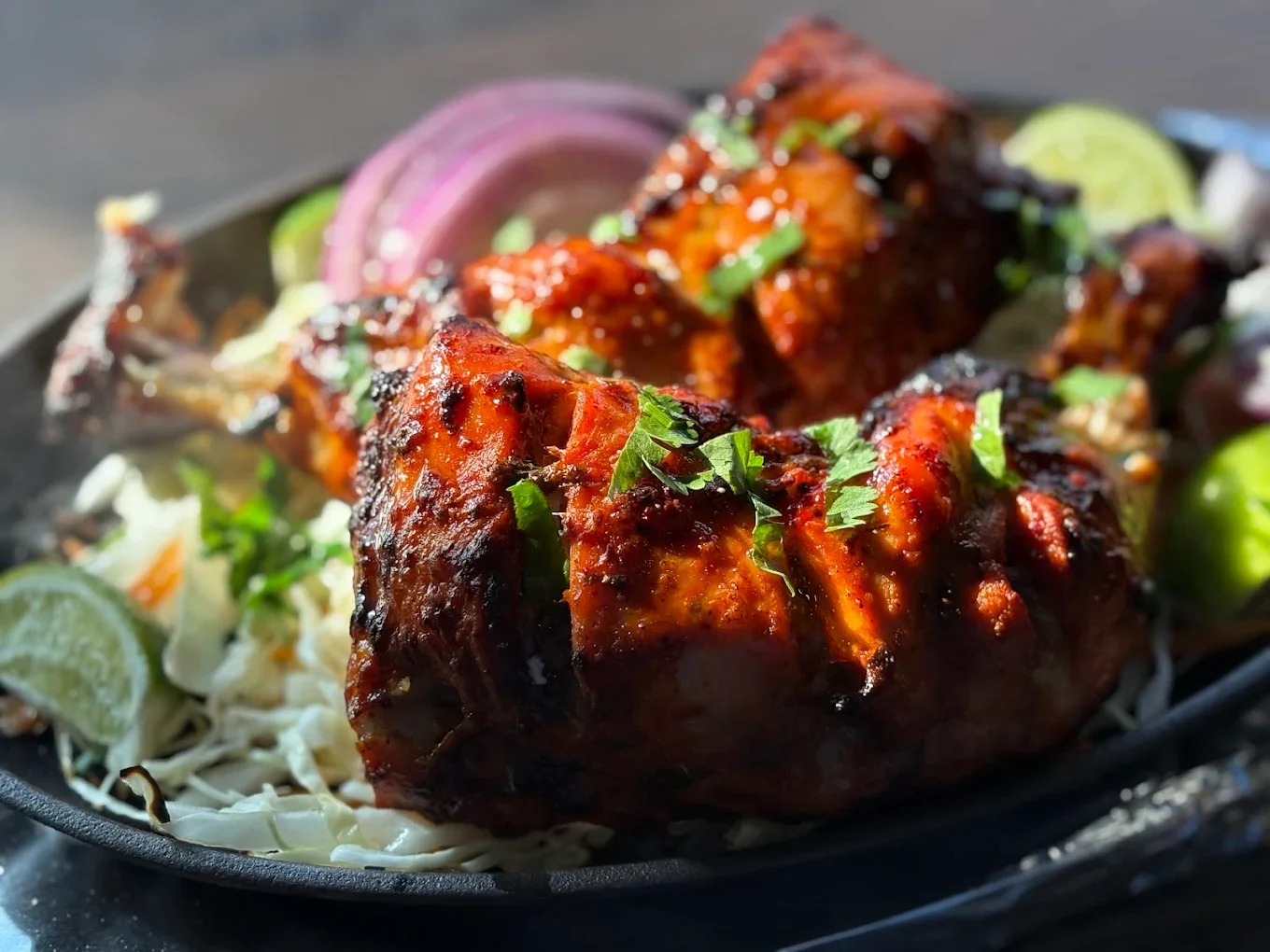 Close-up of spicy grilled chicken drumsticks garnished with cilantro, served with shredded cabbage, purple onion slices, lime wedges, and other fresh vegetables.