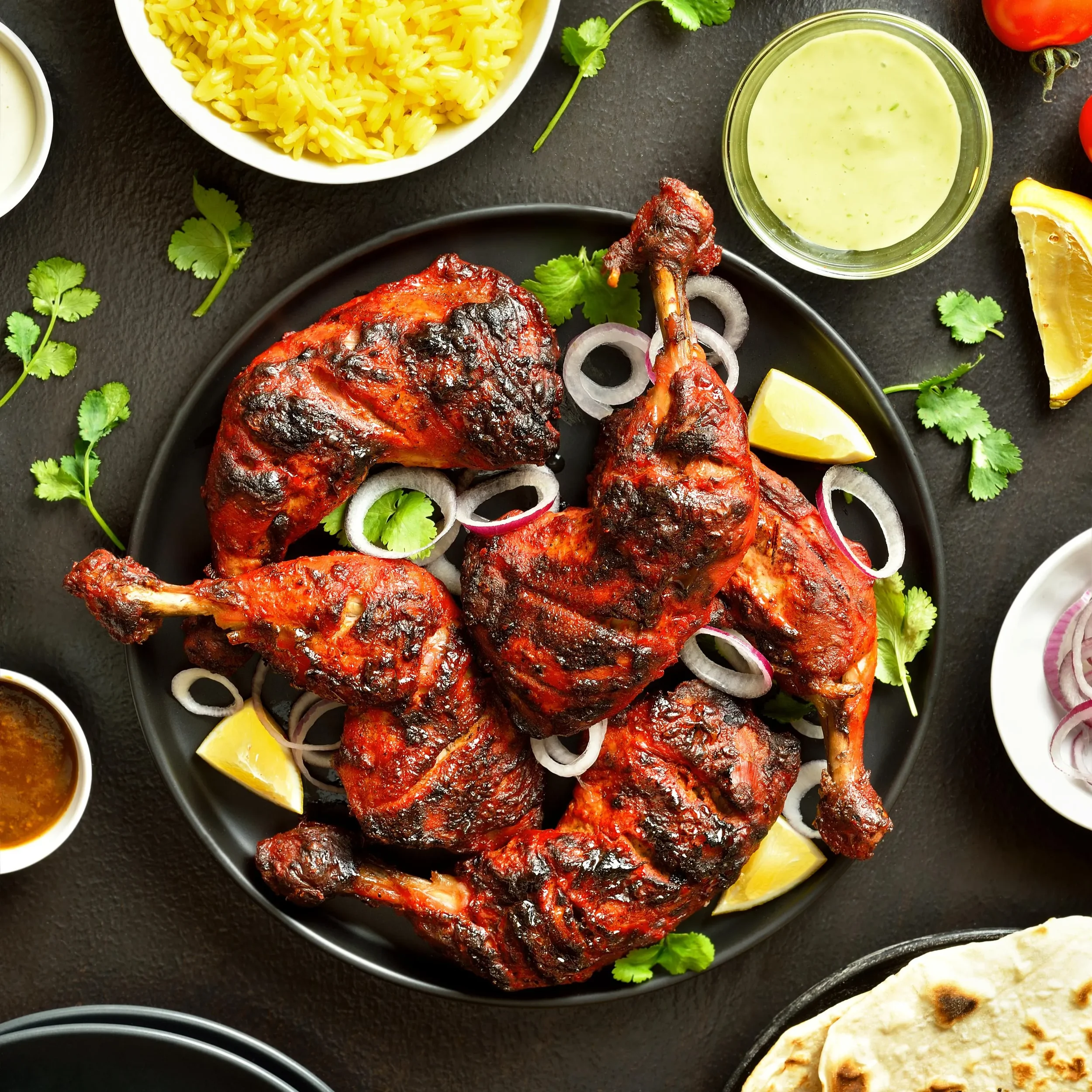 A black plate with four grilled tandoori chicken pieces garnished with sliced onions, cilantro, and lemon wedges, surrounded by bowls of rice, green sauce, and condiments on a black surface.