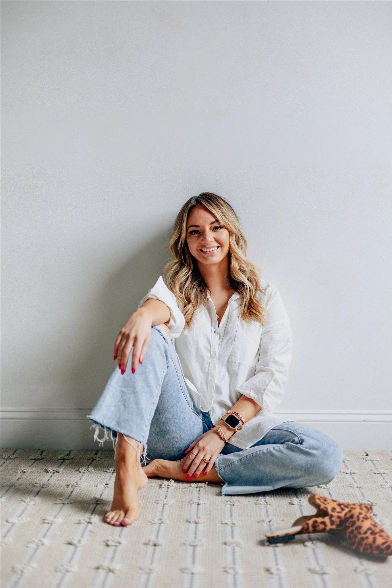 Elle Darcy sitting on the floor against a white wall, smiling, wearing a white shirt and ripped jeans, with a leopard print high heel shoe next to her.