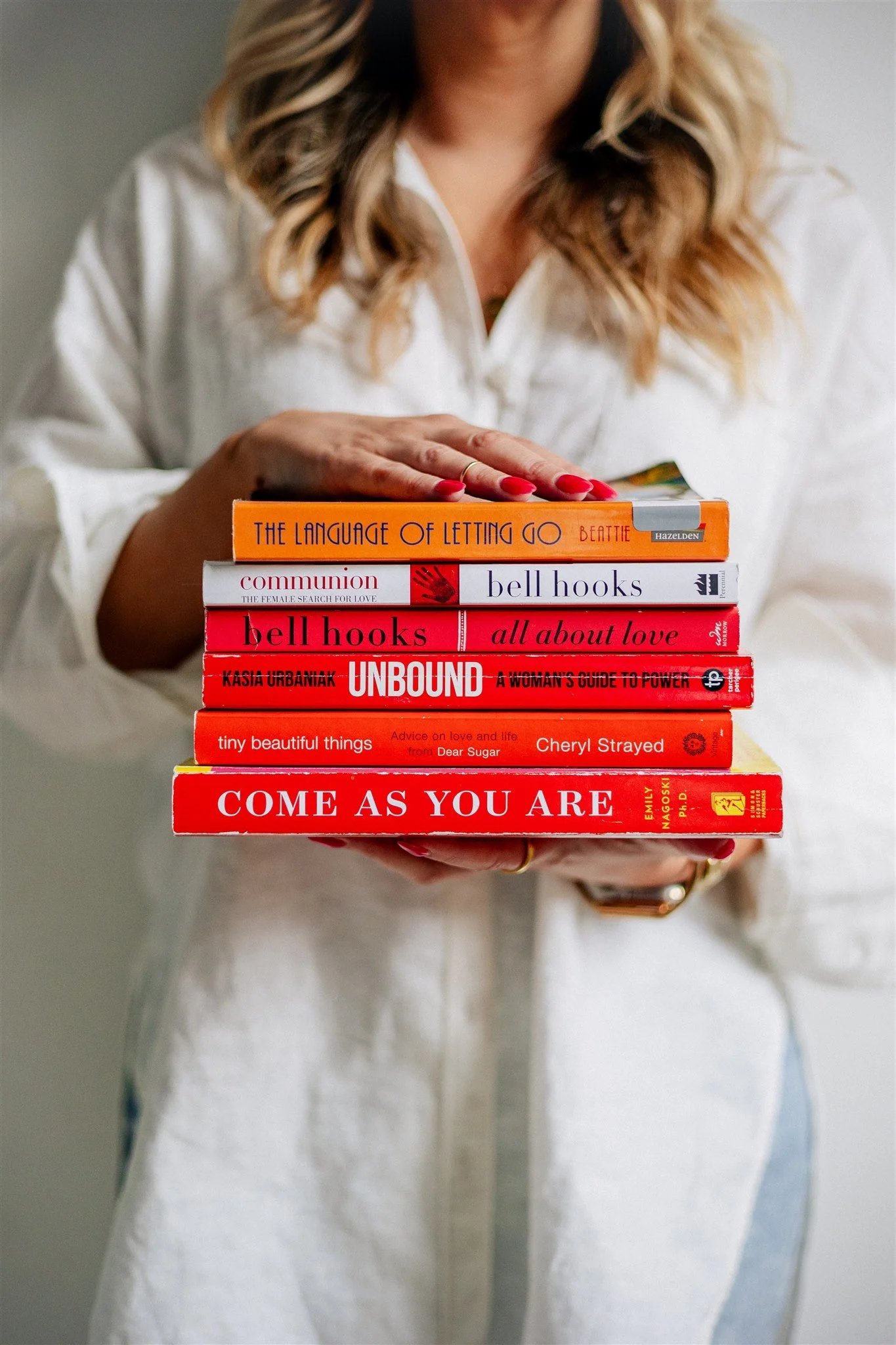 Elle Darcy with blonde, wavy hair is holding a stack of books titled 'The Language of Letting Go,' 'Communion,' 'Bell Hooks: All About Love,' 'Unbound,' 'Tiny Beautiful Things,' and 'Come As You Are.'