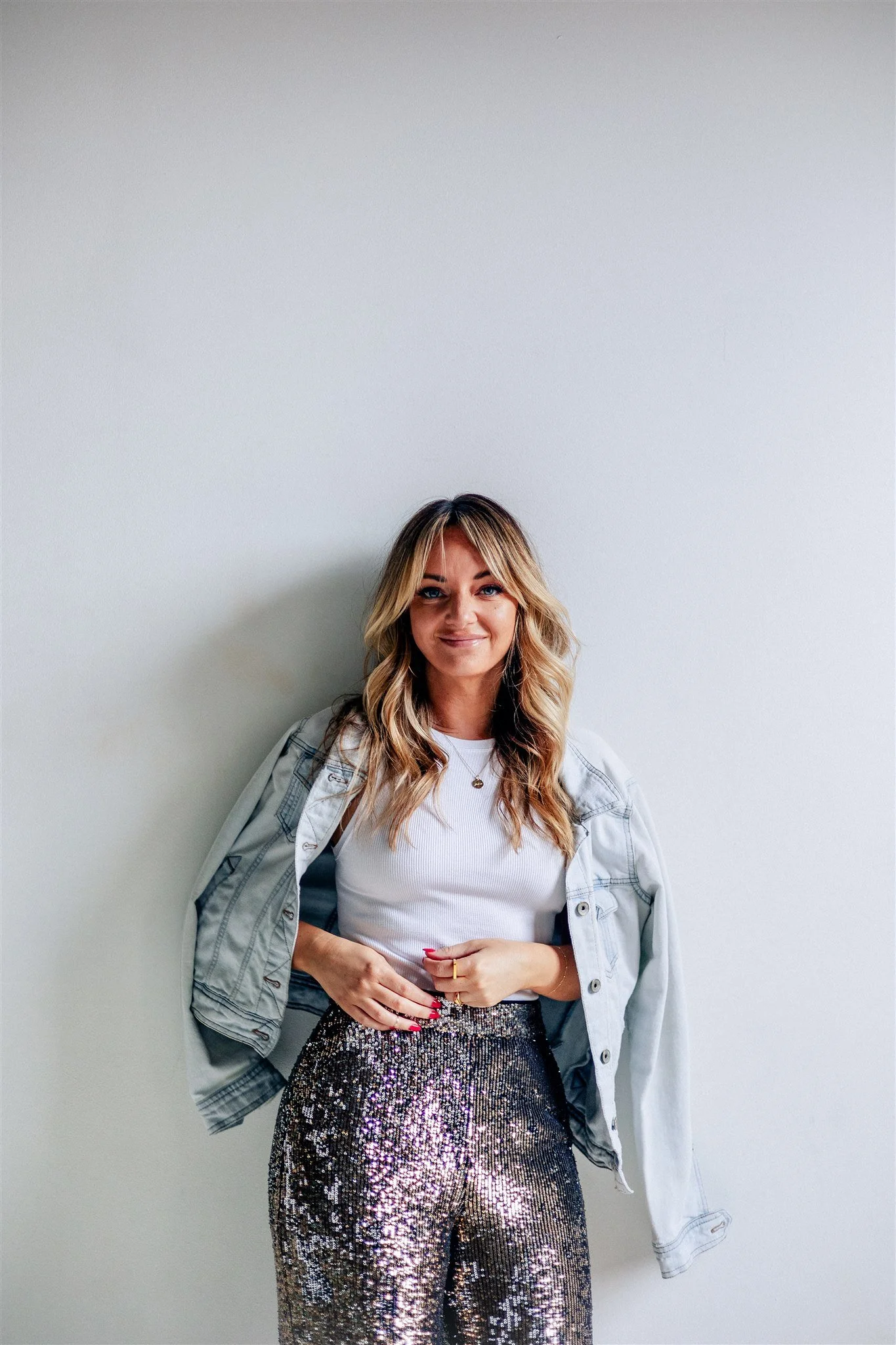 Elle Darcy with blonde, wavy hair wearing a white top, a light denim jacket draped over her shoulders, and high-waisted, shiny, sequined pants standing against a plain white wall.