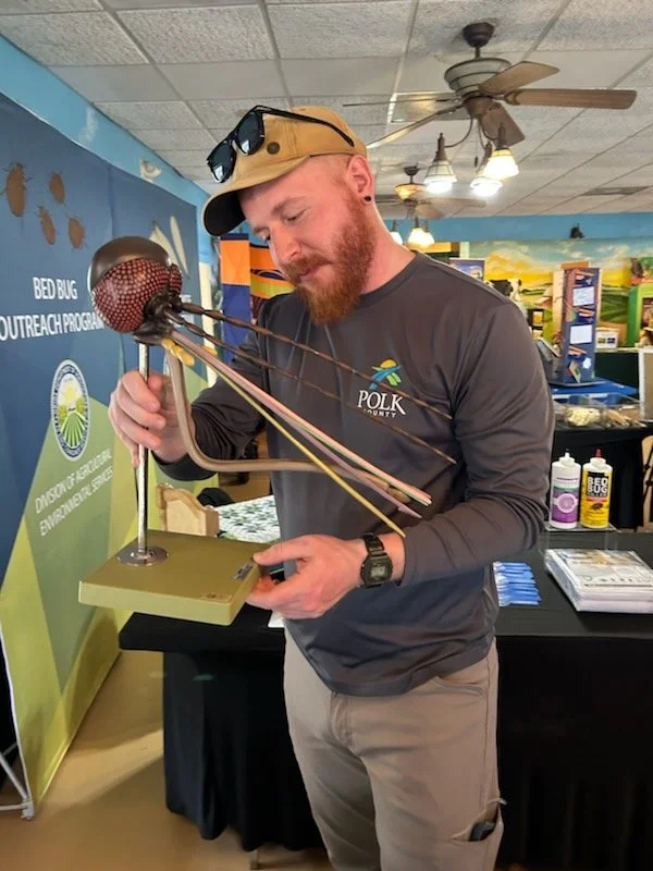 A man with a beard and sunglasses on his cap is examining a model of a bed bug made of plastic and metal at an indoor educational display. He is wearing a dark gray long sleeve shirt and beige pants.