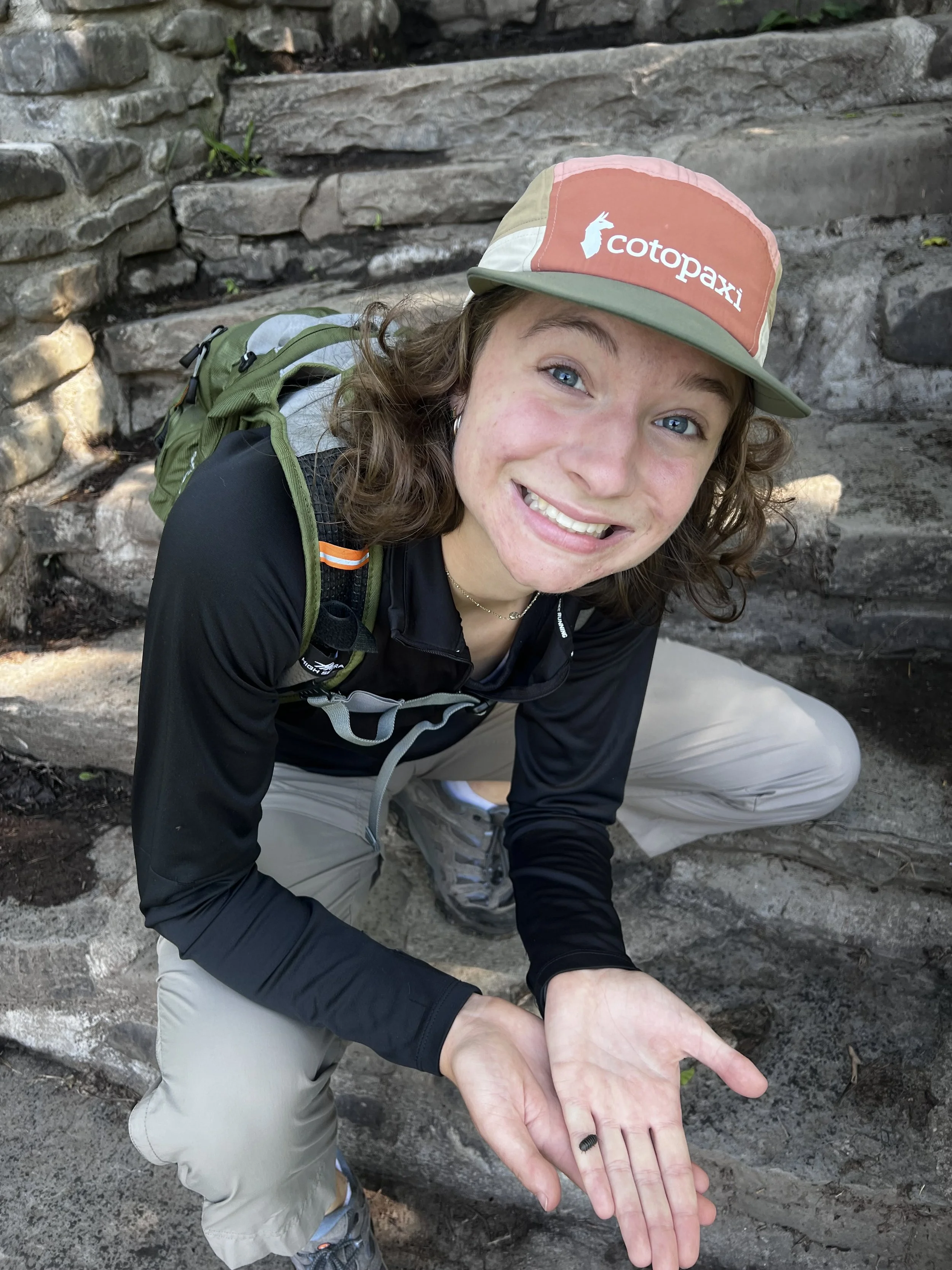 A person wearing a Cotopaxi hat, black jacket, light gray pants, and hiking shoes, holding a small black object in their hand while squatting on a stone staircase outdoors.