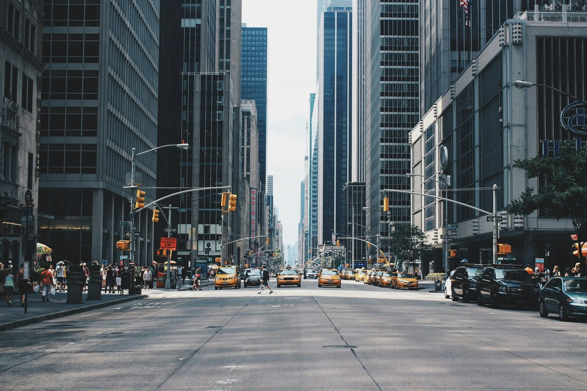 A busy city street lined with tall glass buildings on both sides. Taxis and cars are parked along the curb, and pedestrians are crossing the street. Traffic lights and street signs are visible at the intersection.