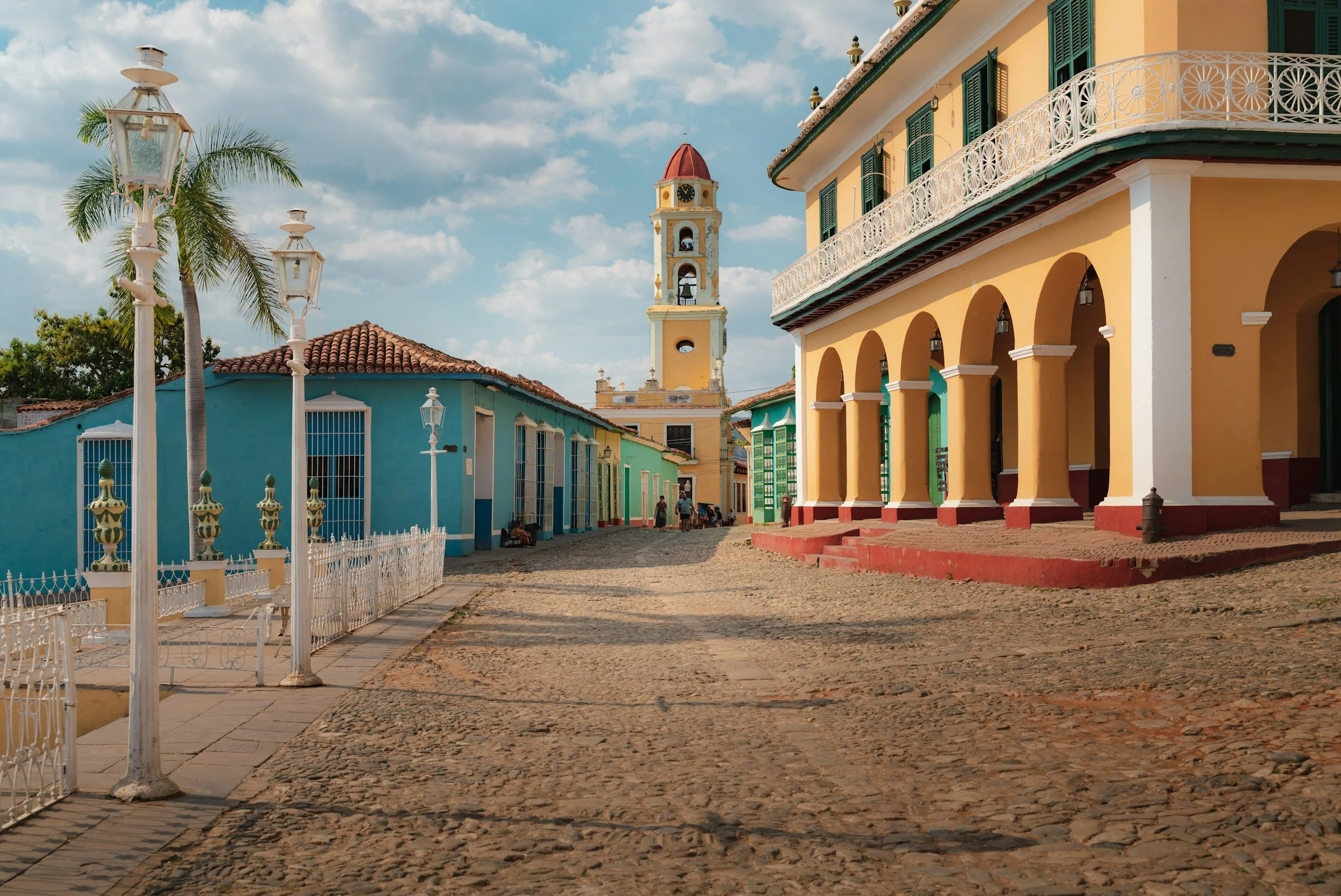 A colorful street scene with pastel yellow and blue buildings, a clock tower with a red dome, palm trees, and vintage street lamps under a partly cloudy sky in a historic town.
