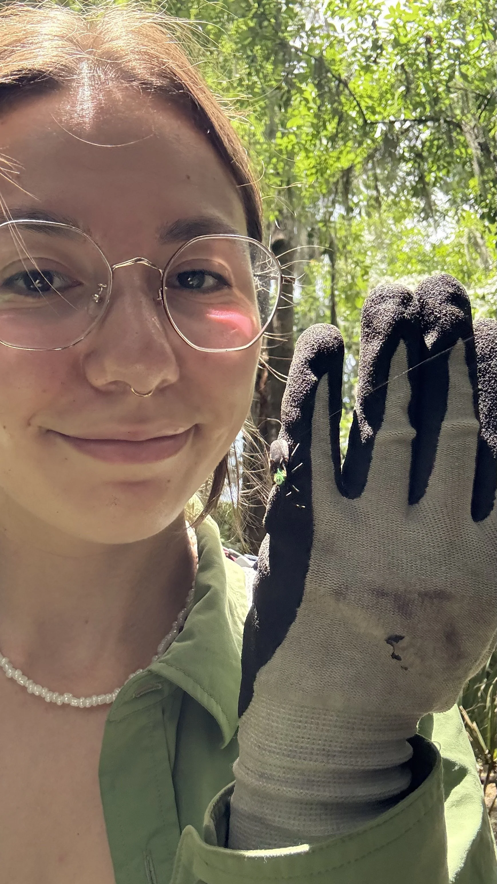 A woman is outdoors in a wooded area, holding a tool or glove, smiling at the camera, wearing glasses, a green jacket, and a pearl necklace.