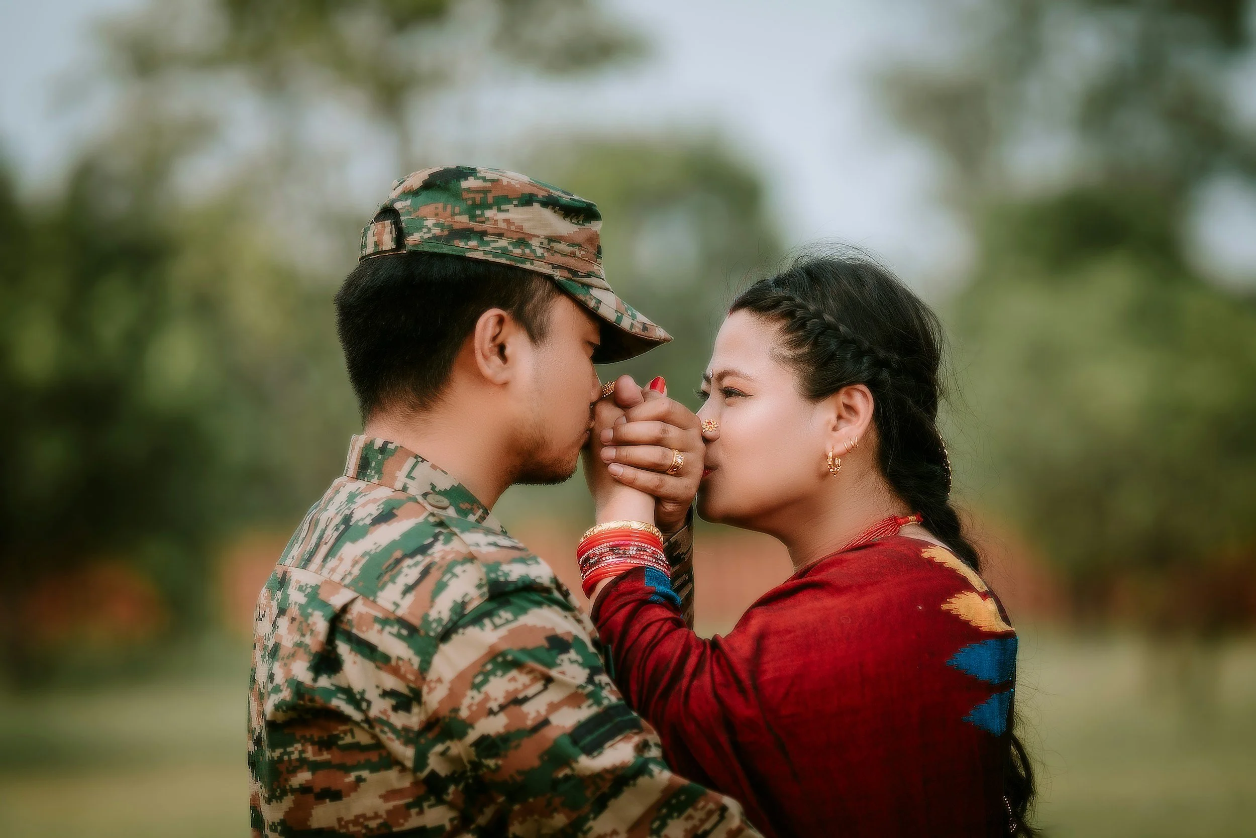 A couple sharing a close, intimate moment outdoors. The man is dressed in a camouflage military uniform, and the woman is wearing traditional Indian clothing with jewelry. They are touching foreheads and holding hands.