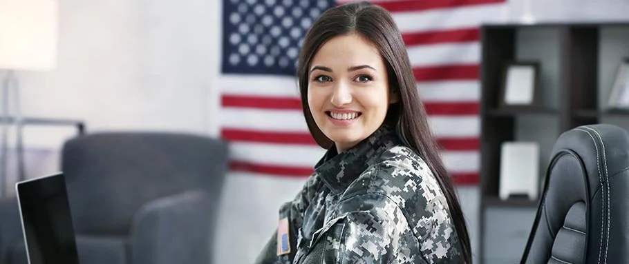 A young woman in military uniform smiling in an office with American flag in background.