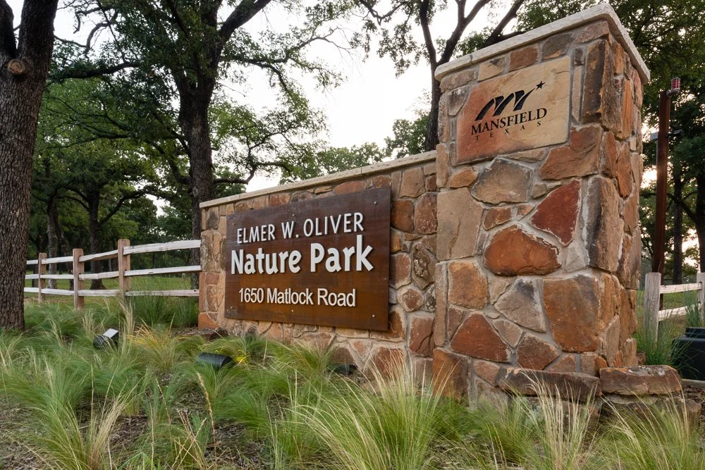 Stone entrance sign for Elmer W. Oliver Nature Park on Matlock Road in Mansfield, Texas, with trees and a white fence in the background.