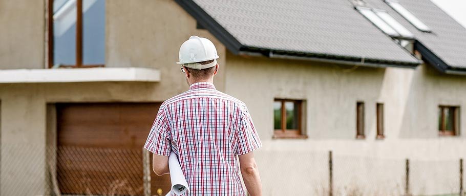 A man wearing a white construction helmet and a red plaid shirt walking in front of a house under construction.