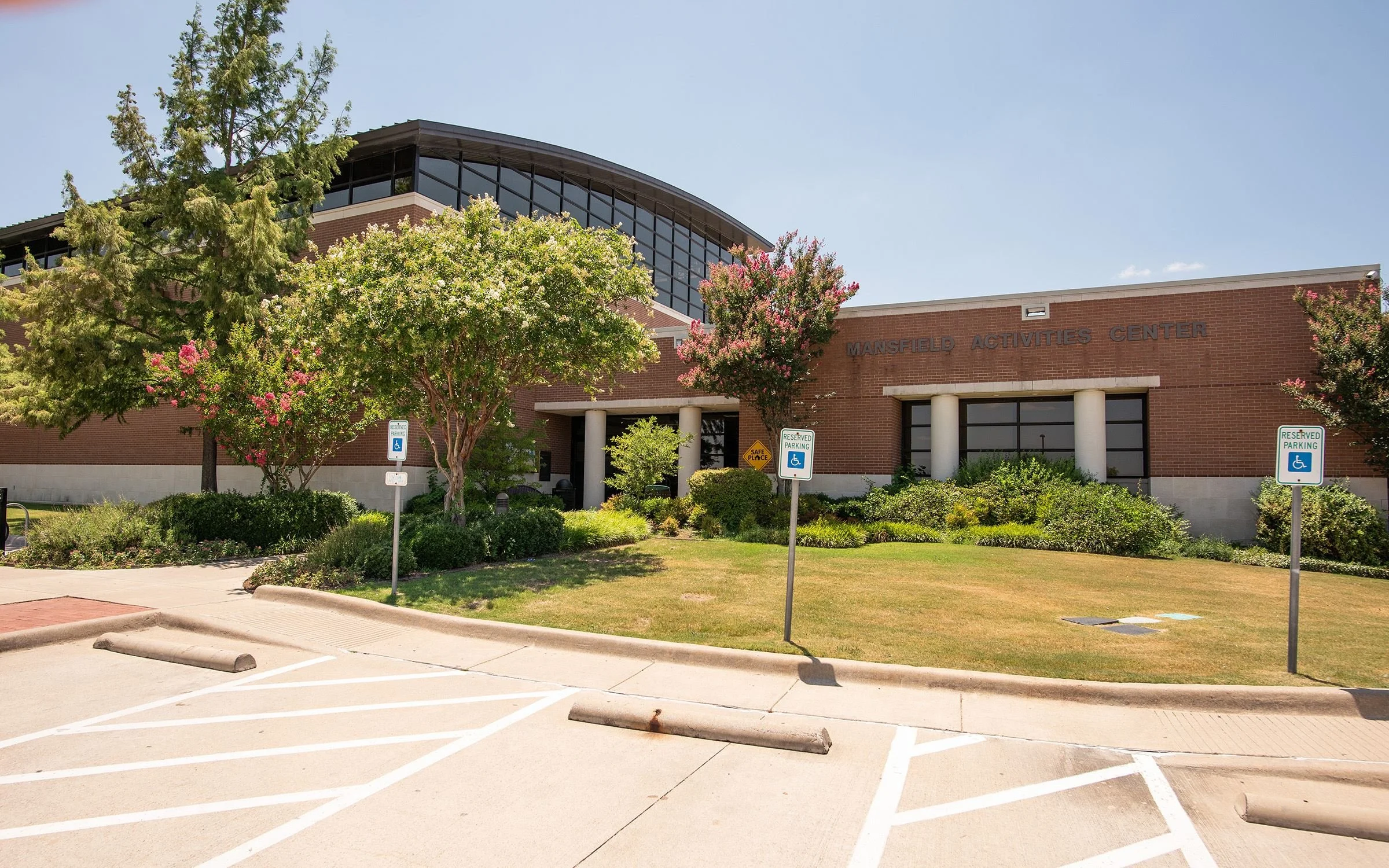 A view of Mansfield Activities Center with trees and reserved parking signs in the foreground, a parking lot with marked spaces, and a building with large windows and a brick exterior under a clear blue sky.