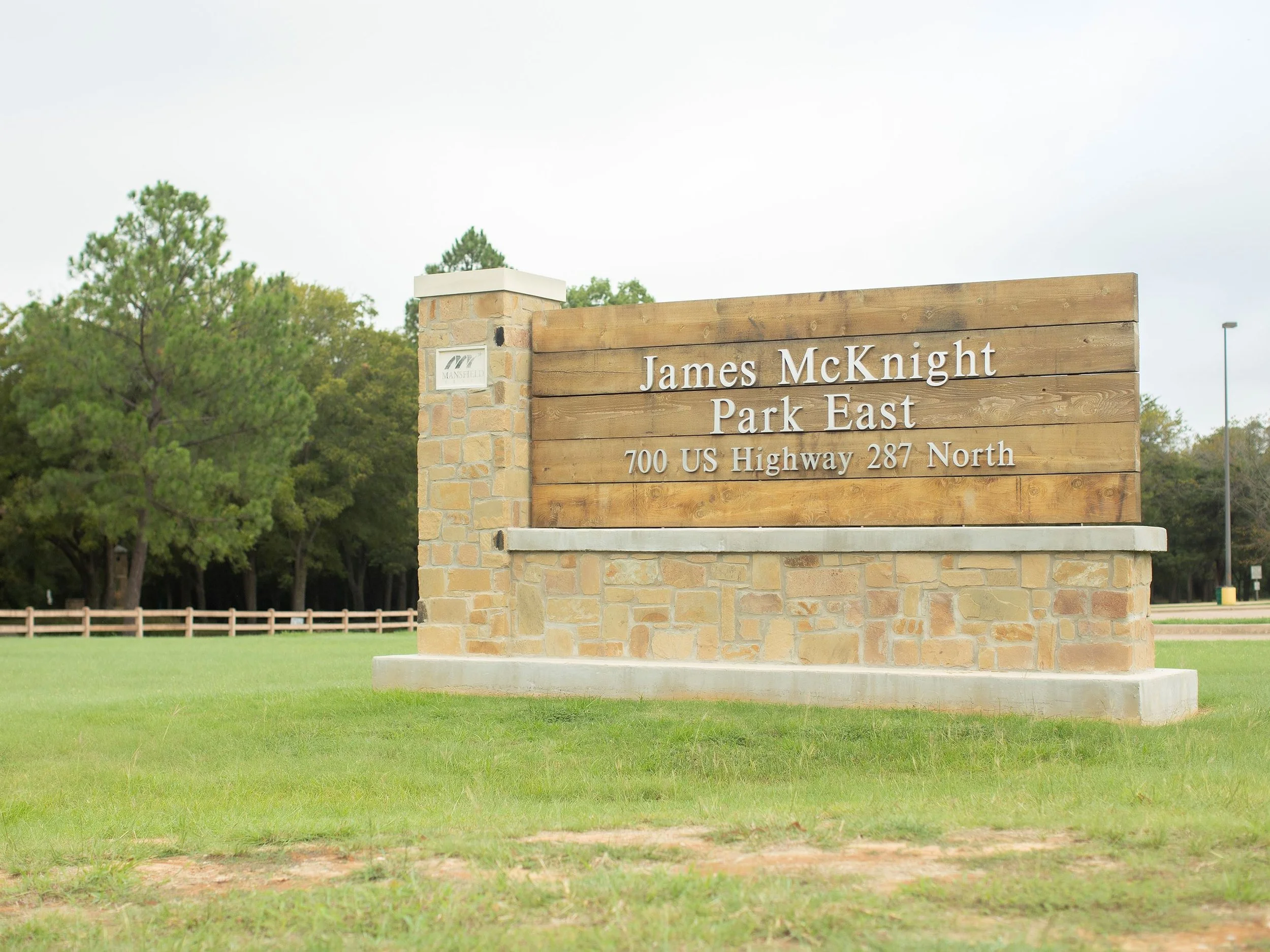 A scenic entrance sign for James McKnight Park East, located at 700 US Highway 287 North, made of wood and stone with a green grassy area in the foreground and trees in the background.