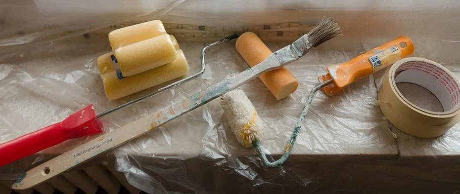 Collection of painting tools including paint rollers, brushes, foam rollers, tape, and a paint tray on a plastic-covered surface.