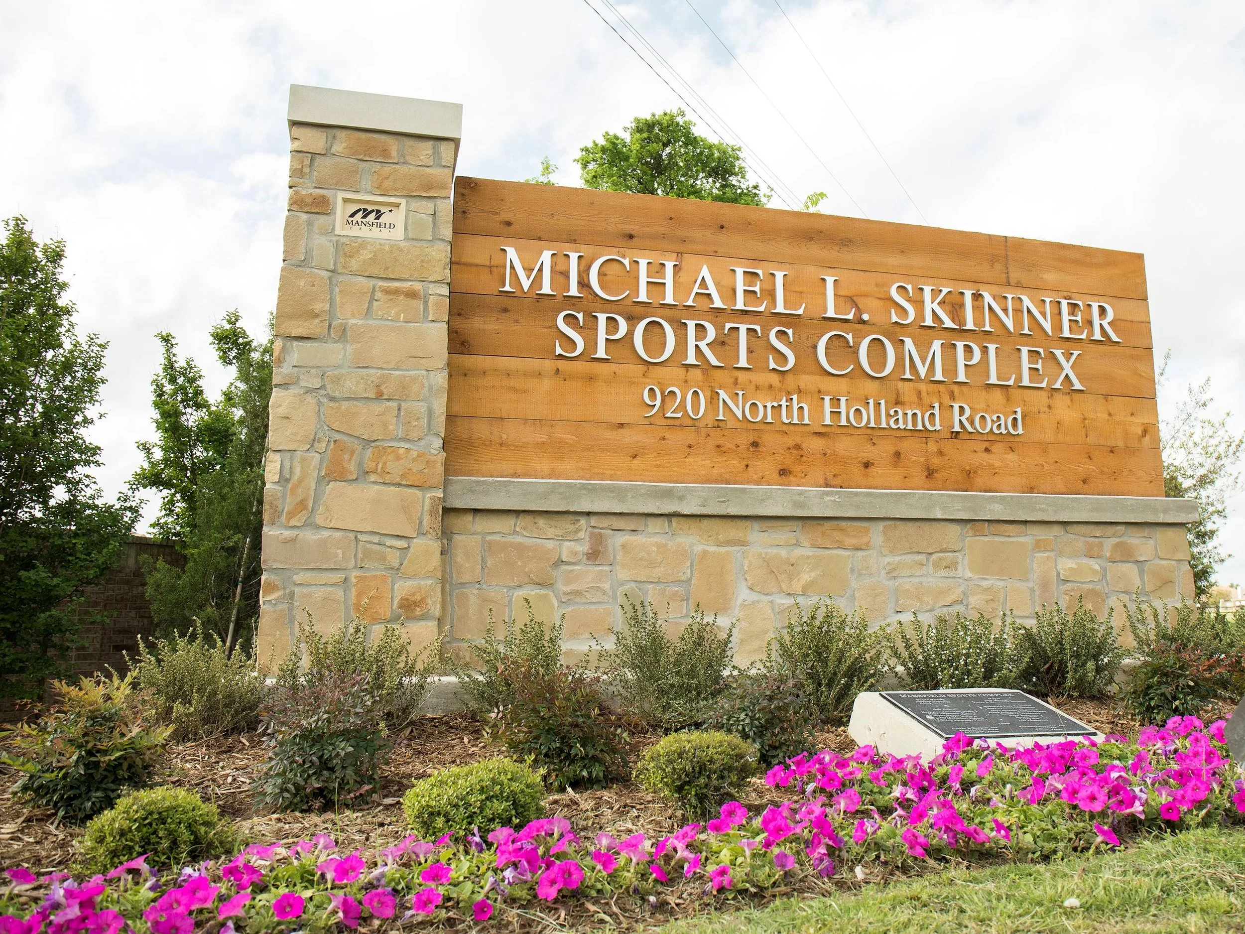Sign for Michael L. Skinner Sports Complex located at 920 North Holland Road, with landscaped flowers and plants in foreground, trees and partly cloudy sky in background.