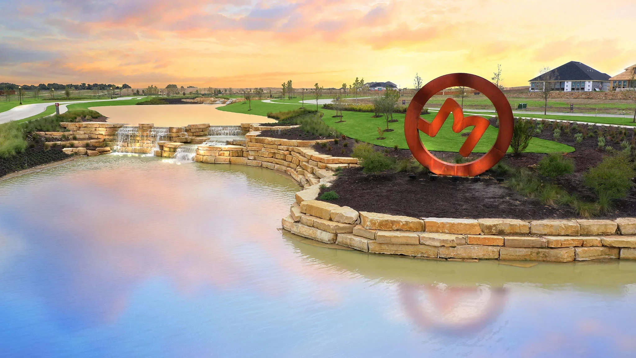 A landscaped park with a small waterfall and pond, a winding sidewalk, young trees, a large orange art sculpture of a heart and medical pulse, and houses in the background during a sunset.