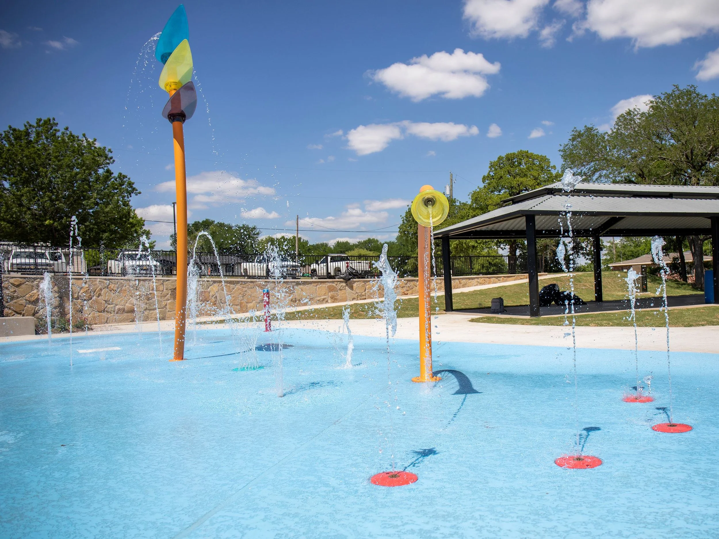 A water park with water spraying from multiple colorful fountains, including orange, yellow, and blue, on a bright sunny day with a partly cloudy sky. There is a pavilion and trees in the background.