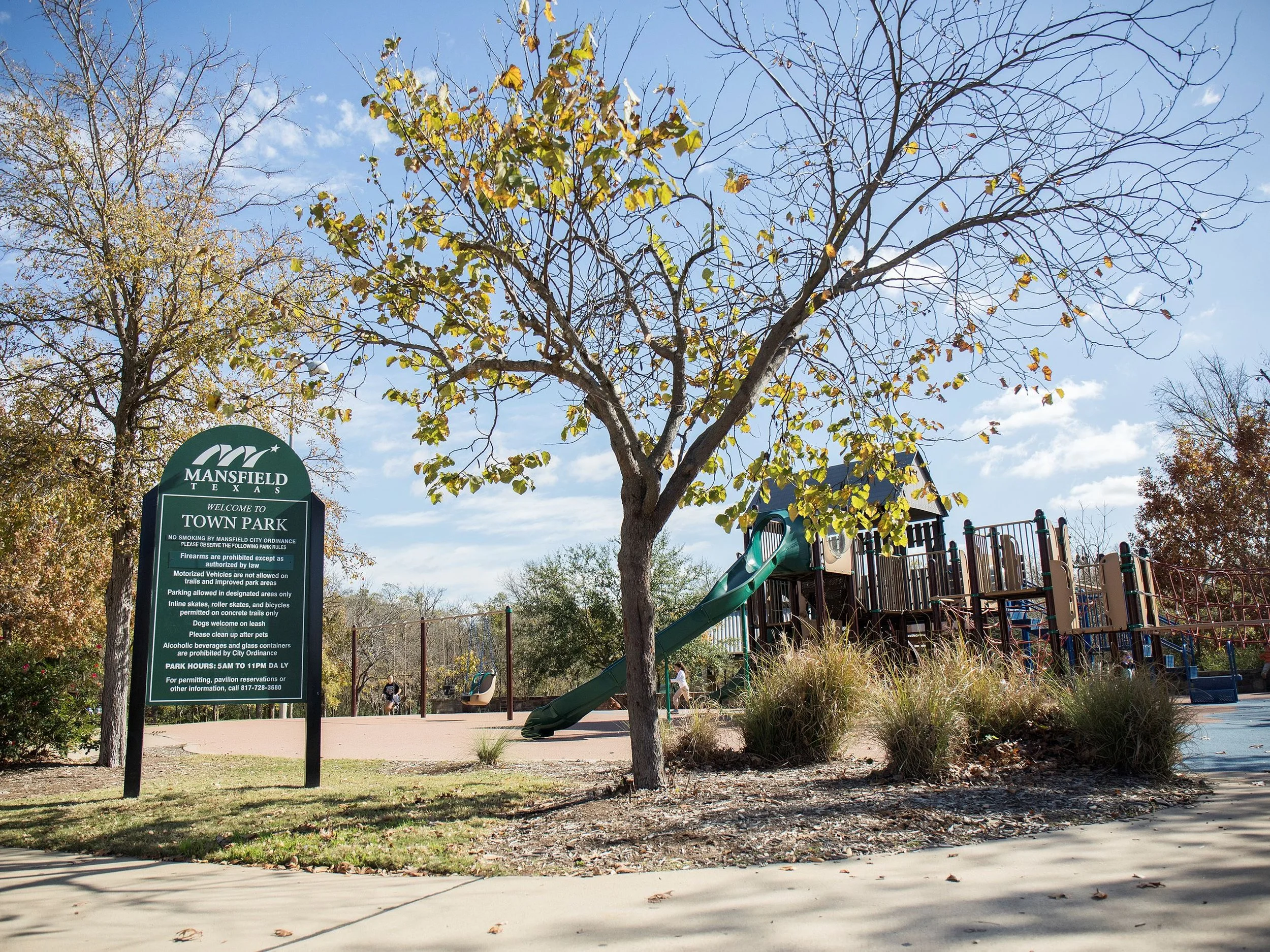 A park scene with a playground and a sign reading 'Mansfield Texas Welcome to Town Park.' The playground has a slide, trees with some leaves and some without, and kids playing. The sky is clear with some clouds.