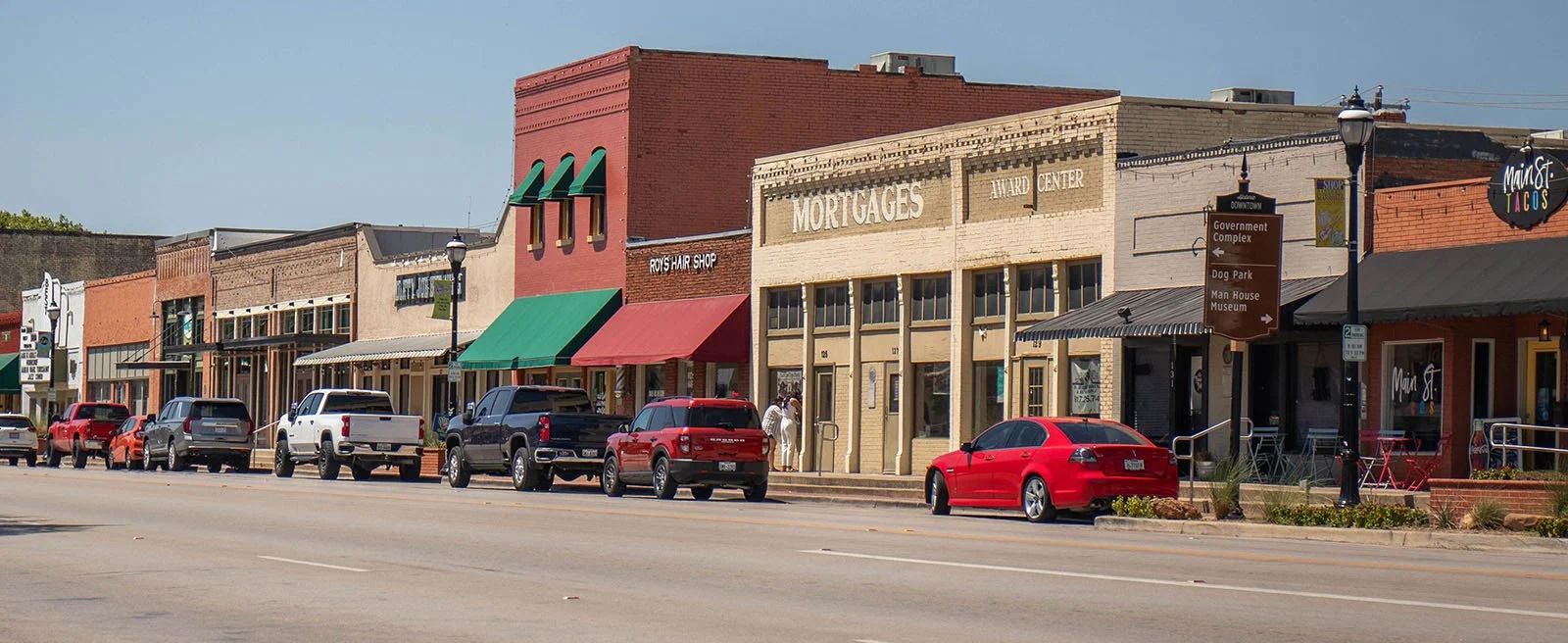 Main street view with historic brick buildings, parked cars, and shops, including a hair salon and a mortgage office, under clear skies.