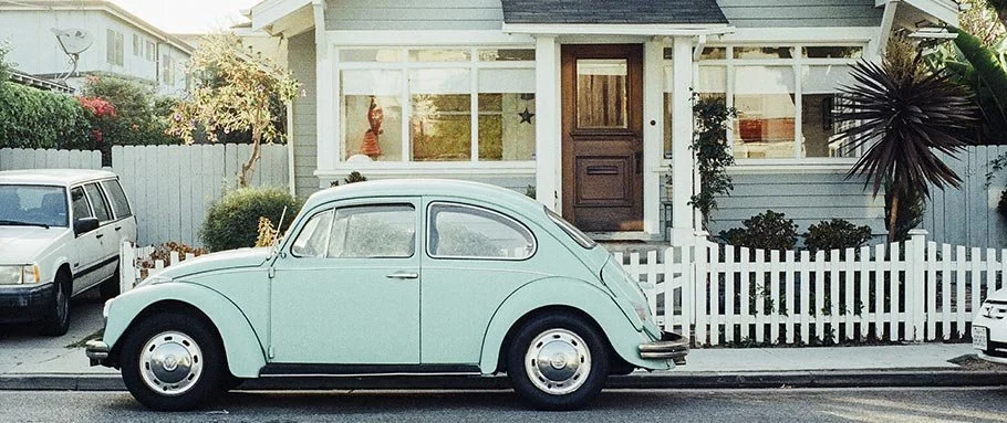 A vintage light blue Volkswagen Beetle parked in front of a house with a white picket fence.