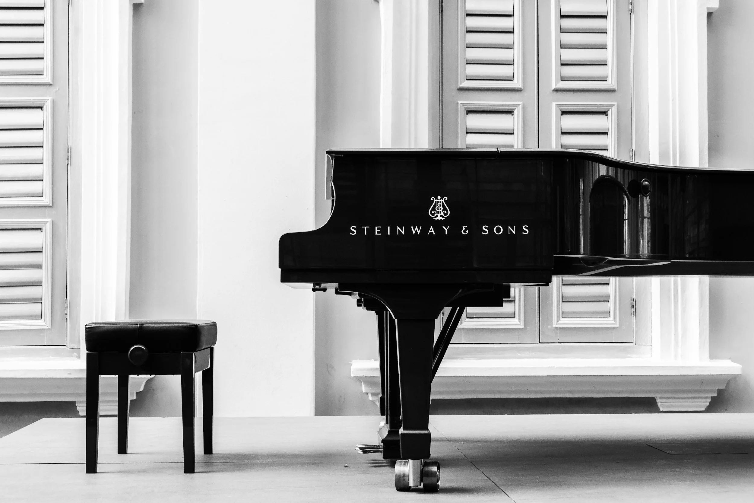 A black Steinway & Sons grand piano on a wooden floor in a room with white walls and closed window shutters.