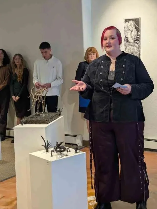 A woman with red hair talking at an art gallery, with three sculptures on display in front of her and a group of people listening in the background.