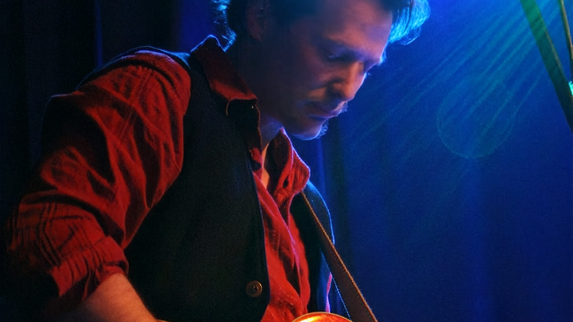 Ray Taylor, a man with brown hair wearing a red plaid shirt and black vest looking down playing guitar in a dimly lit room with blue lighting.