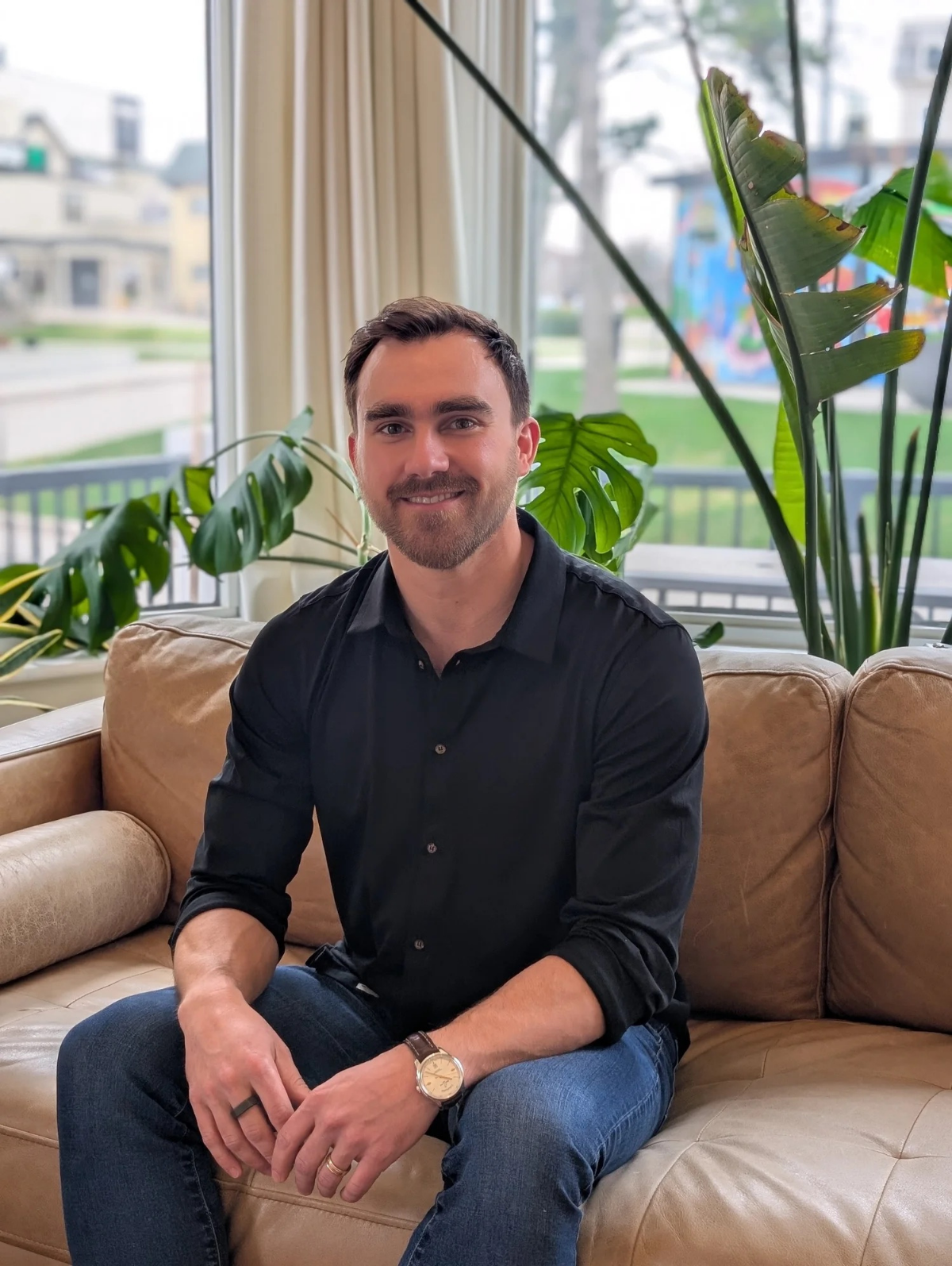Chris Cebulski sitting on a beige leather couch surrounded by large green houseplants, with a window showing a suburban neighborhood outside.