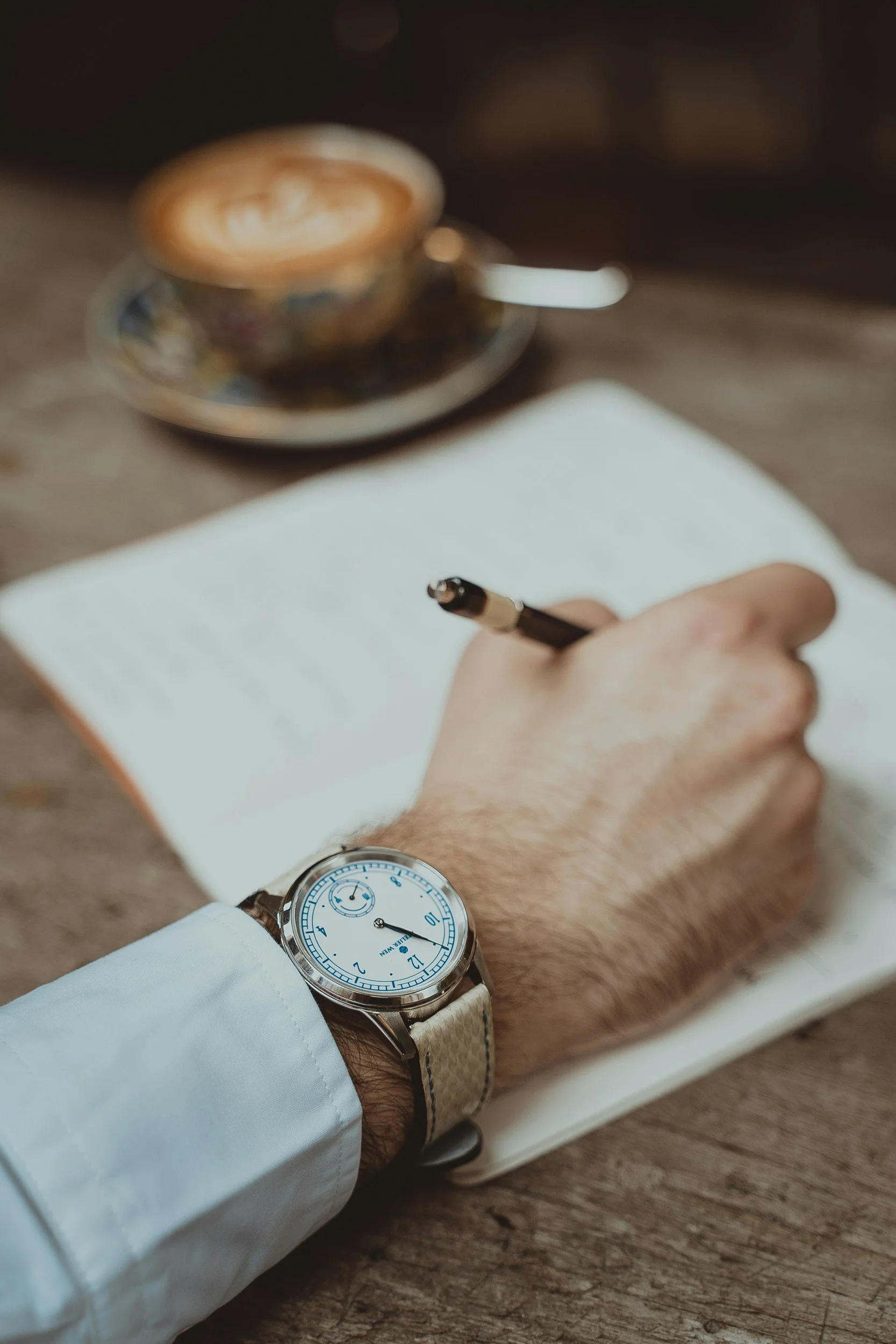 A person wearing a white shirt with a silver watch on their wrist, writing on a notepad with a black pen, resting on a wooden surface, with a cup of coffee and a saucer blurred in the background.