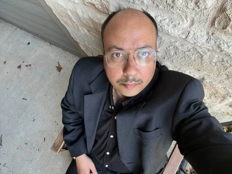 A man in formal attire with glasses taking a selfie outdoors, sitting on a bench next to a stone wall.