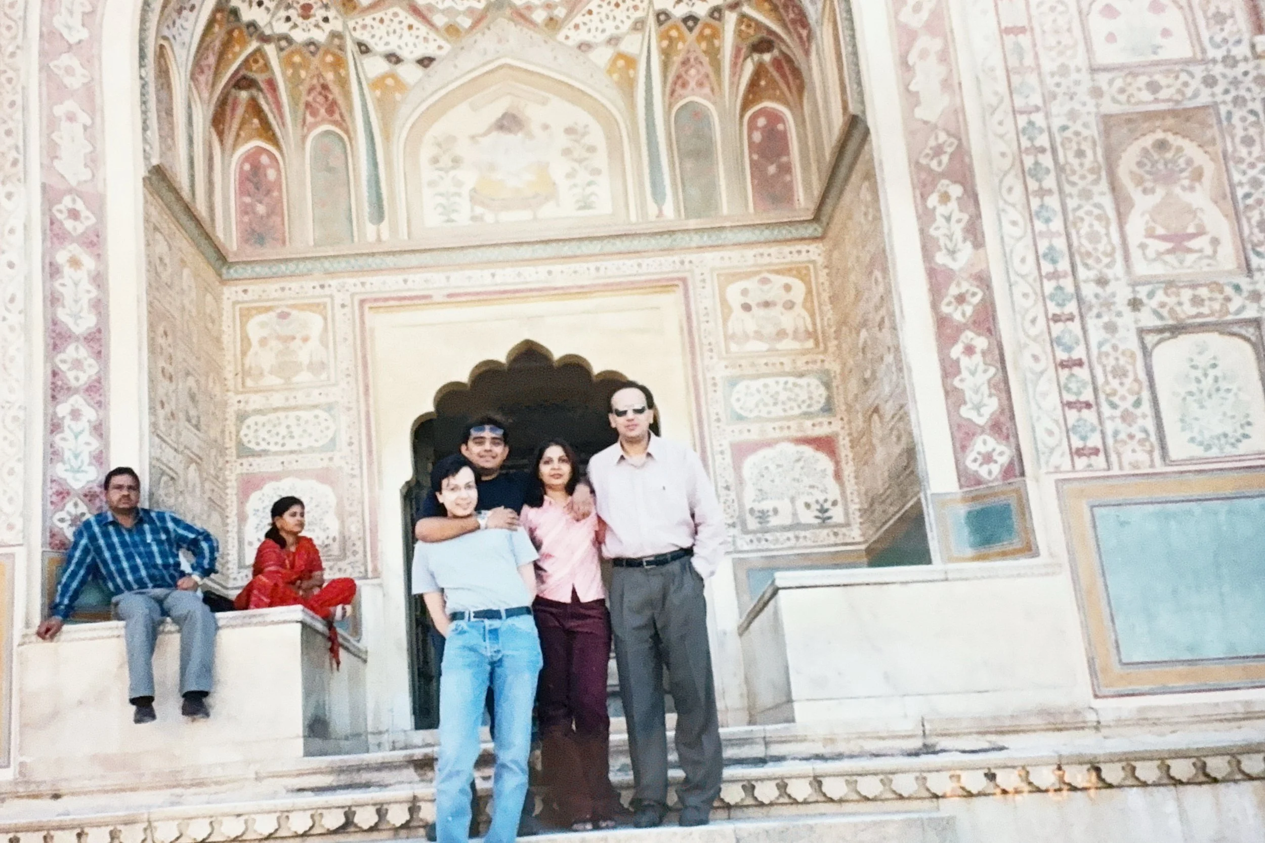 Group of five people in front of a painted palace wall, with two people sitting on ledge to the side.