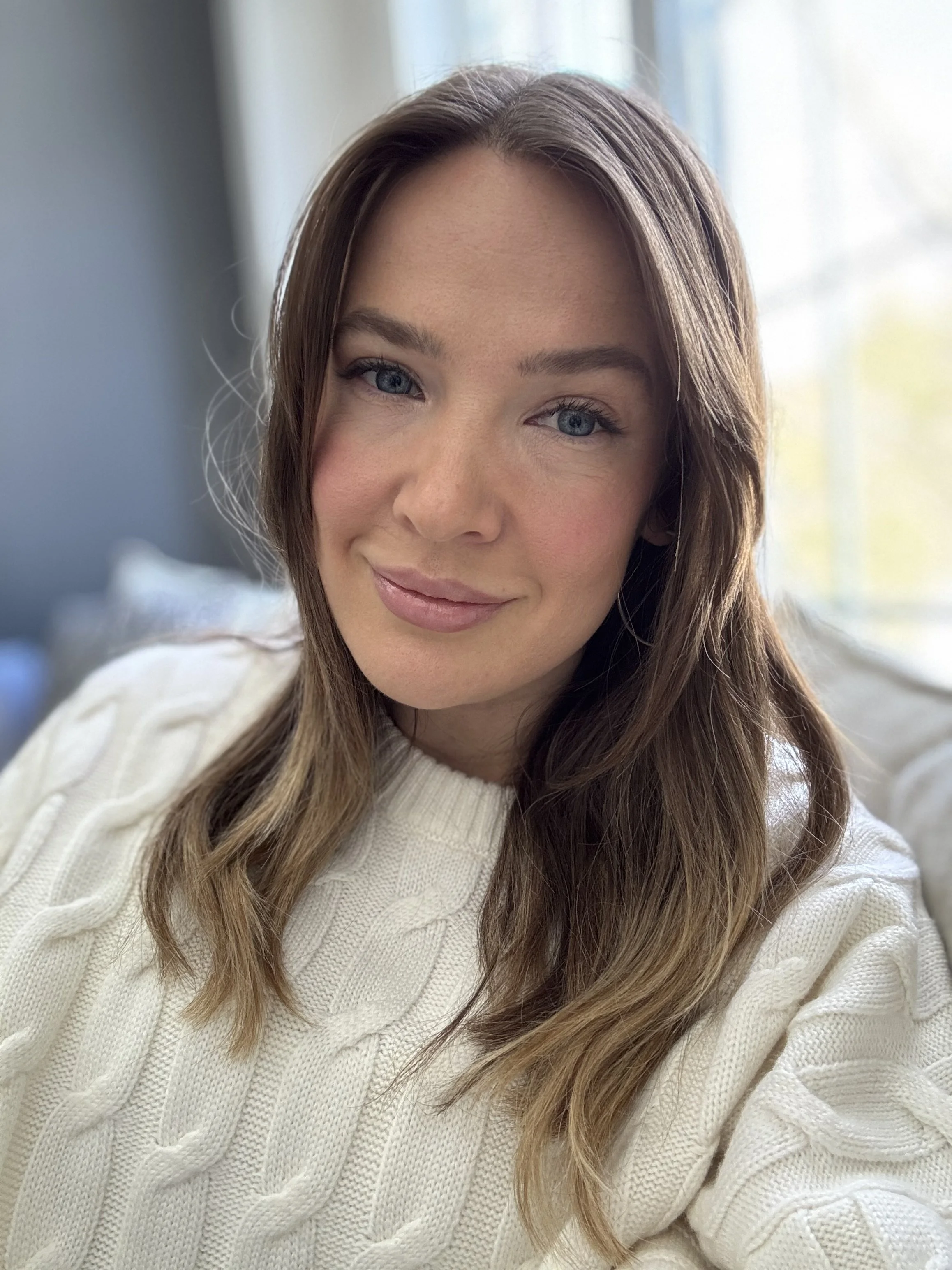 Close-up of a woman with long wavy brown hair, blue eyes, and a light makeup, smiling softly indoors near a window with sheer curtains, wearing a white cable-knit sweater.
