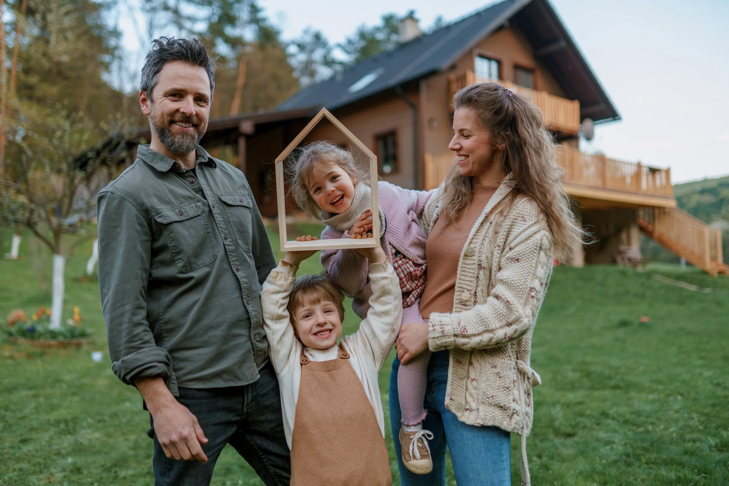 A family of four standing outside in a grassy yard in front of a wooden house, smiling and holding a small wooden house model.