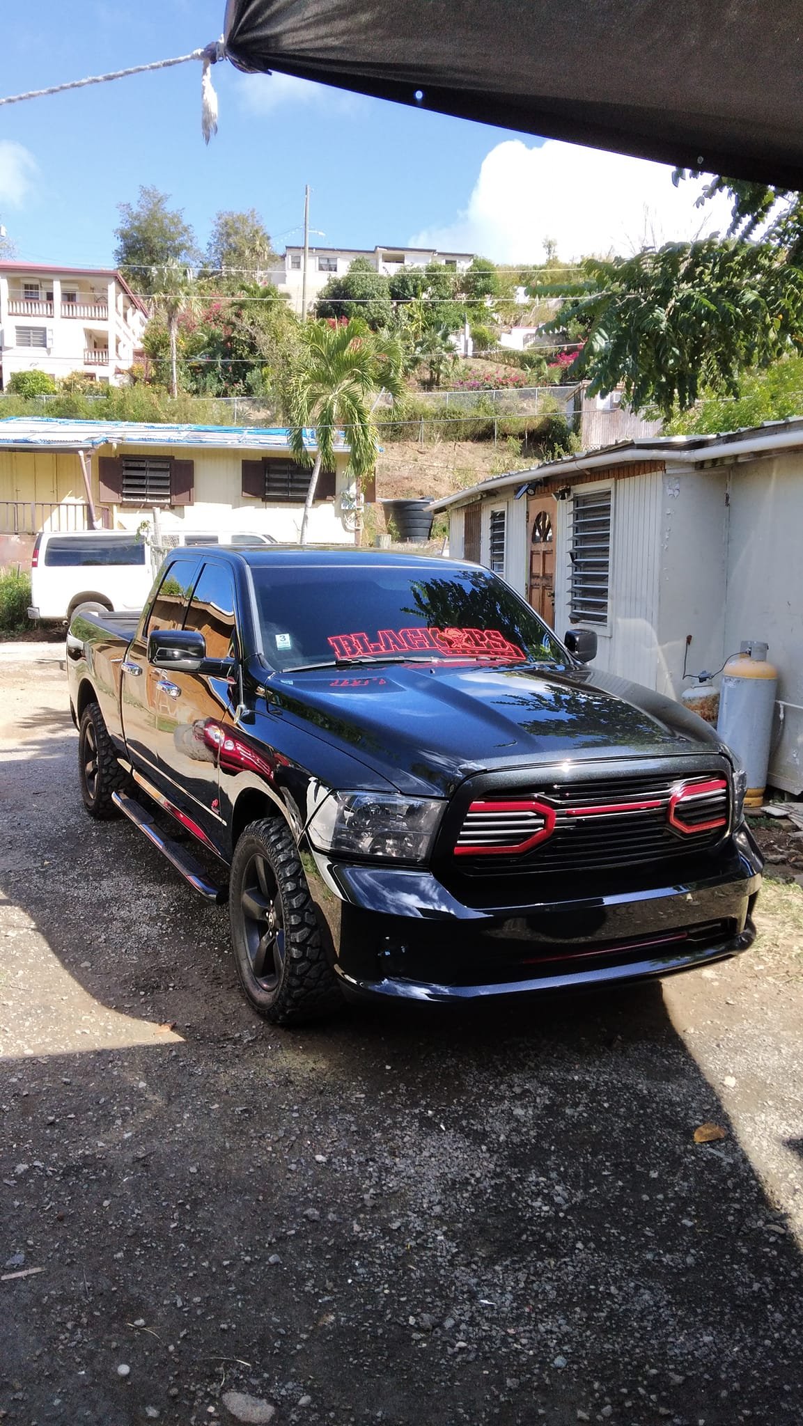 A black pickup truck with red accents and tinted windows parked outdoors on a dirt surface, with a residential hillside and trees in the background.