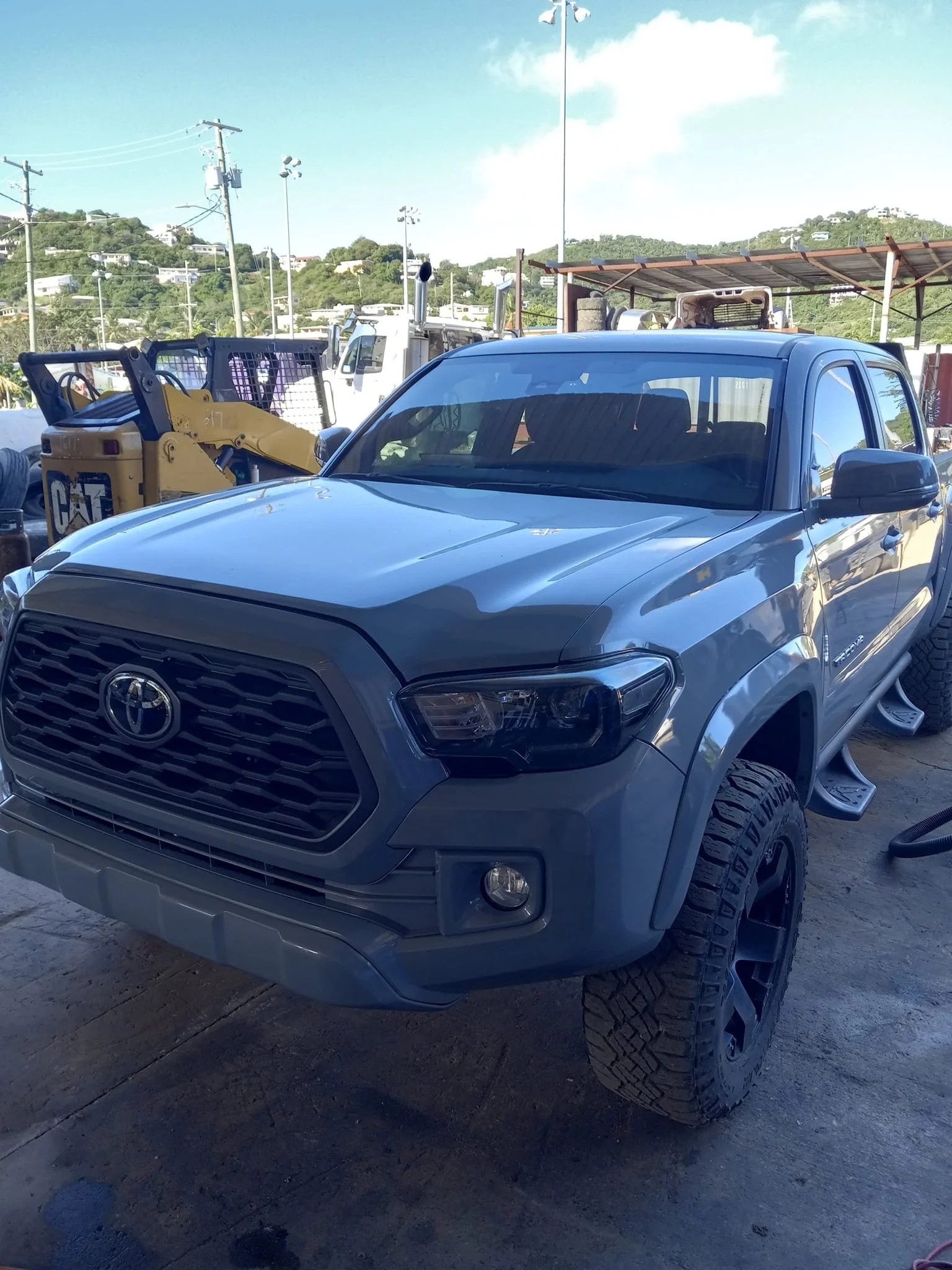 Silver Toyota pickup truck with large tires parked outdoors near construction equipment, with a hill and houses in the background under a blue sky.