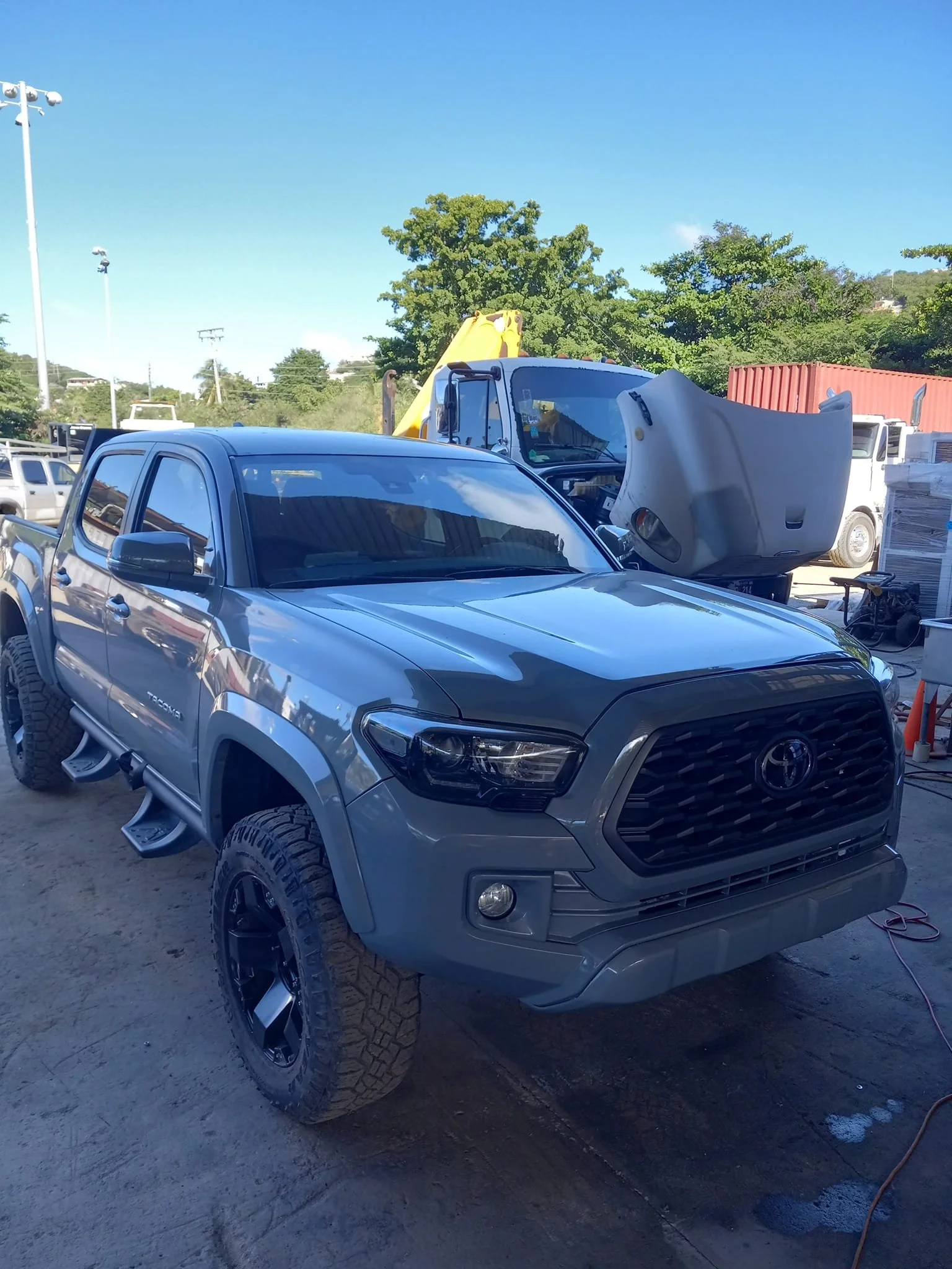A black Toyota Tacoma pickup truck parked on a dirt lot with a gray oversized vehicle and yellow construction equipment behind it, green trees and a blue sky in the background.