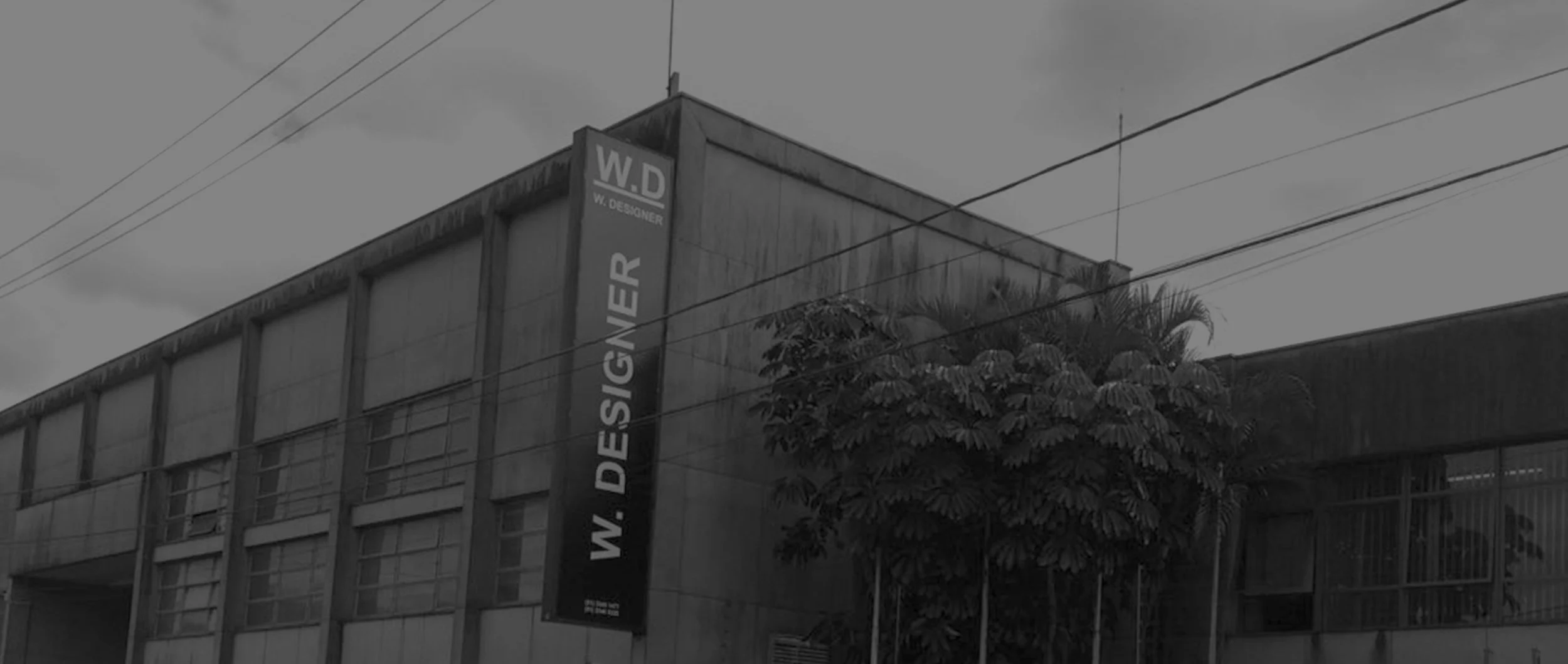 Prédio de concreto com placa de sinalização vertical na fachada, com árvores ao fundo e linhas de energia no céu nublado.