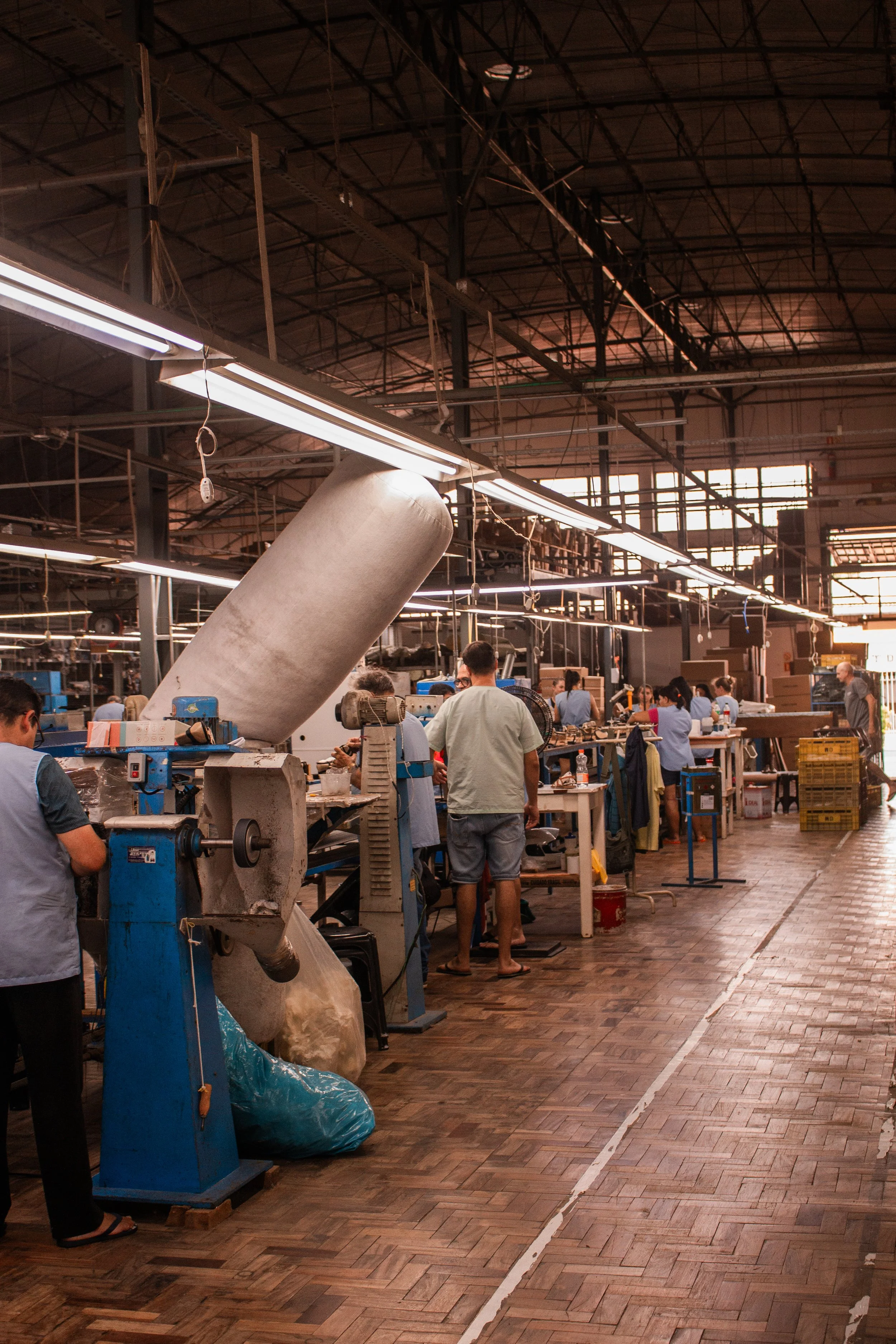 Imagem de uma fábrica com pessoas trabalhando ao longo de várias estações de produção, com máquinas e materiais diversos, iluminação artificial e estrutura metálica no teto.