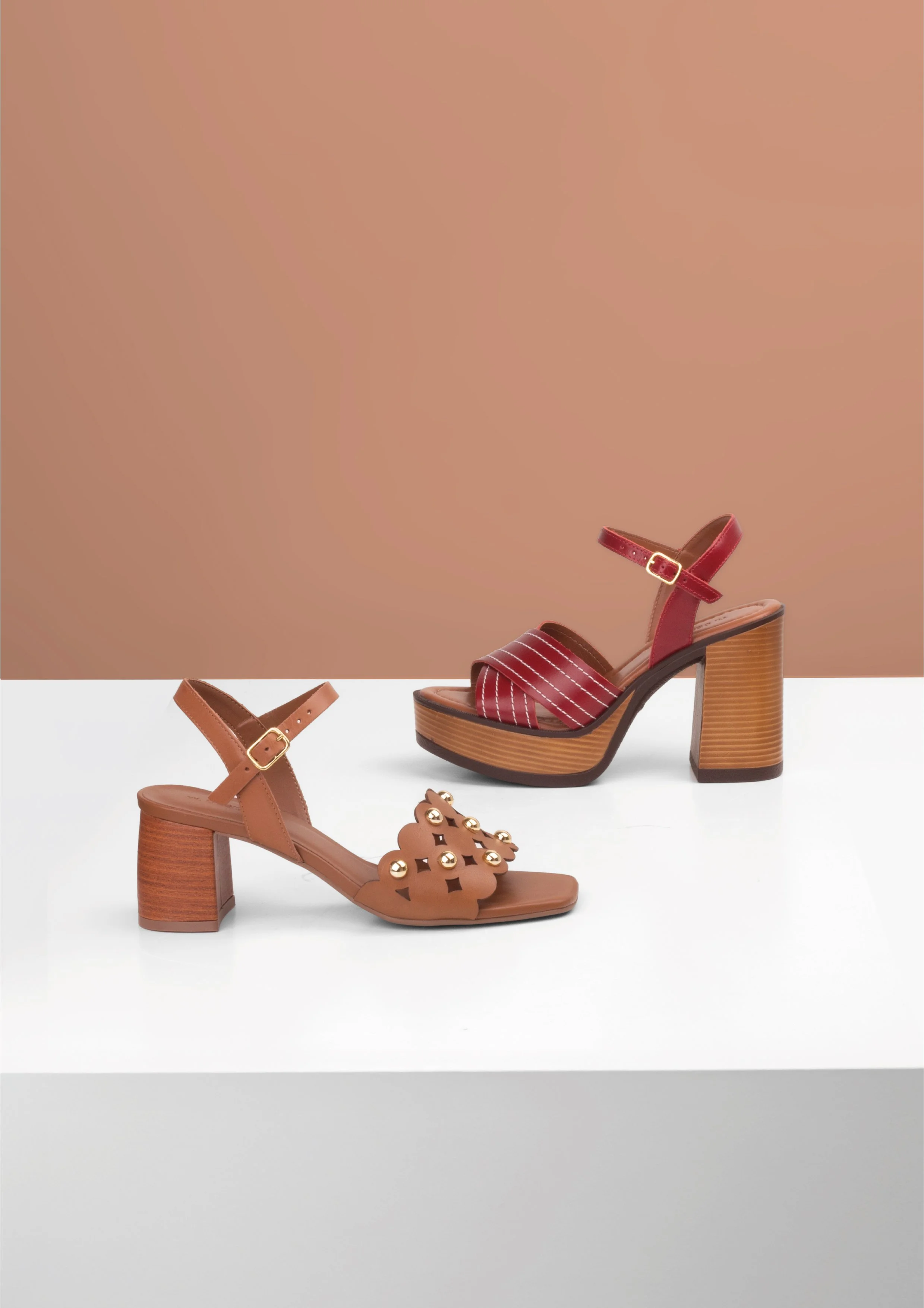 Dois pares de sapatos femininos de salto, um marrom com detalhes decorativos dourados e um vermelho com listras brancas, exibidos sobre uma superfície branca com fundo de cor bege.