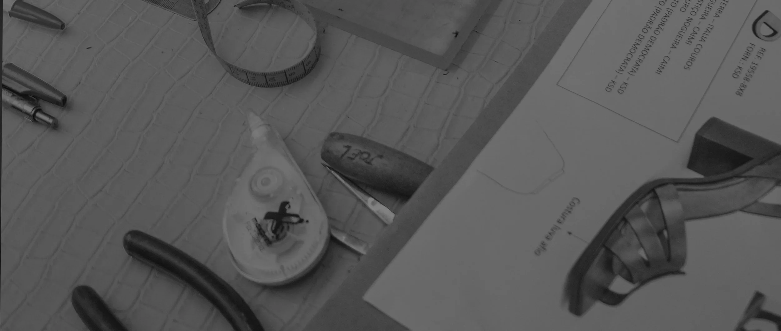Ferramentas e materiais de trabalho sobre uma mesa de vidro com revestimento de azulejos. Inclui uma fita métrica, um tubo de cola, uma lâmina, papel com texto, prancheta, e um par de pinças.