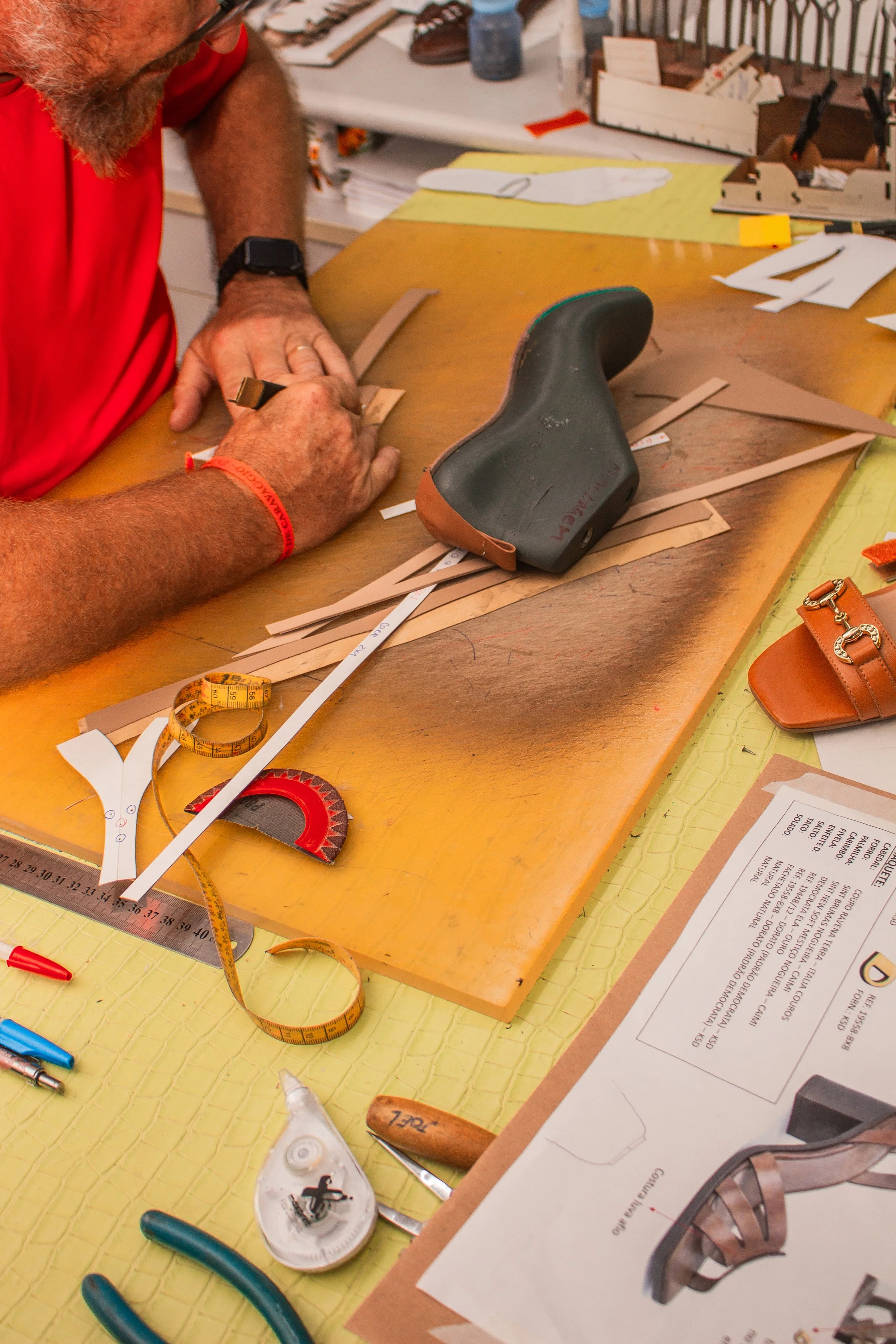 Pessoa trabalhando na confecção de sapatos, cortando ou marcando couro com ferramentas na bancada de trabalho, com materiais de costura e papel de molde ao redor.