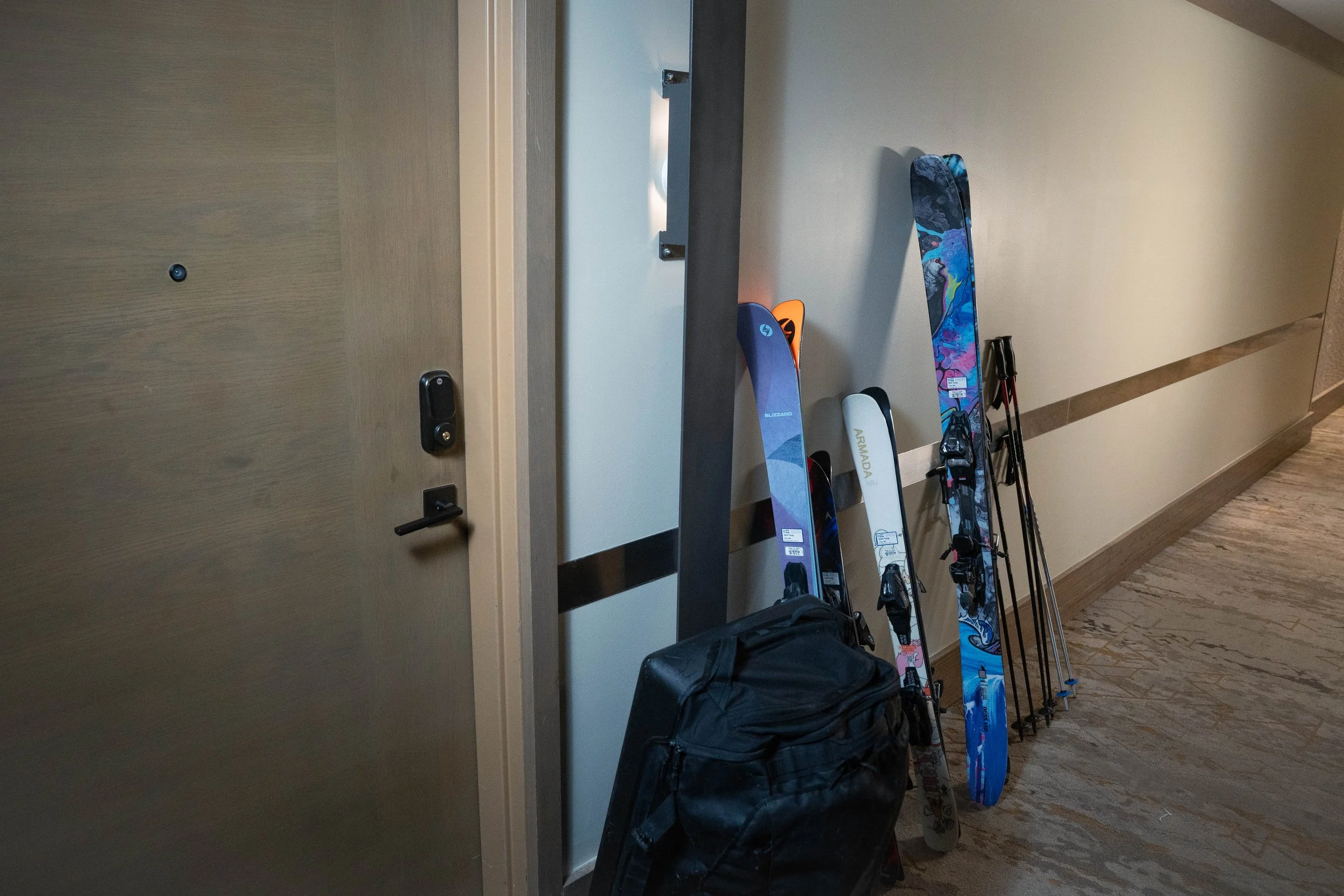 A hotel hallway with a beige wall and wooden door. Leaning against the wall are several skis with colorful designs, and a black backpack is on the floor nearby.