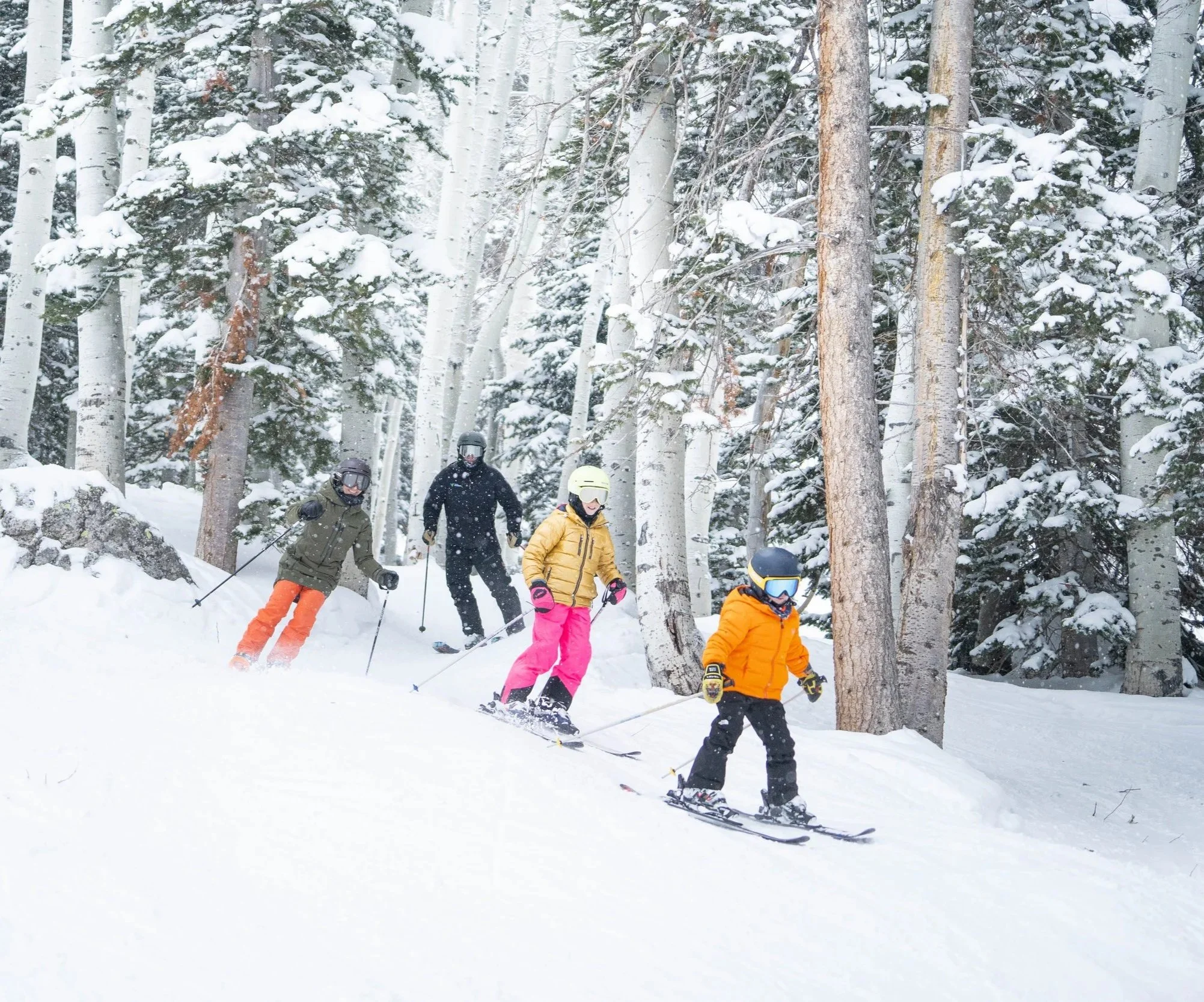A group of five children skiing down a snowy slope in a forested area with snow-covered trees.