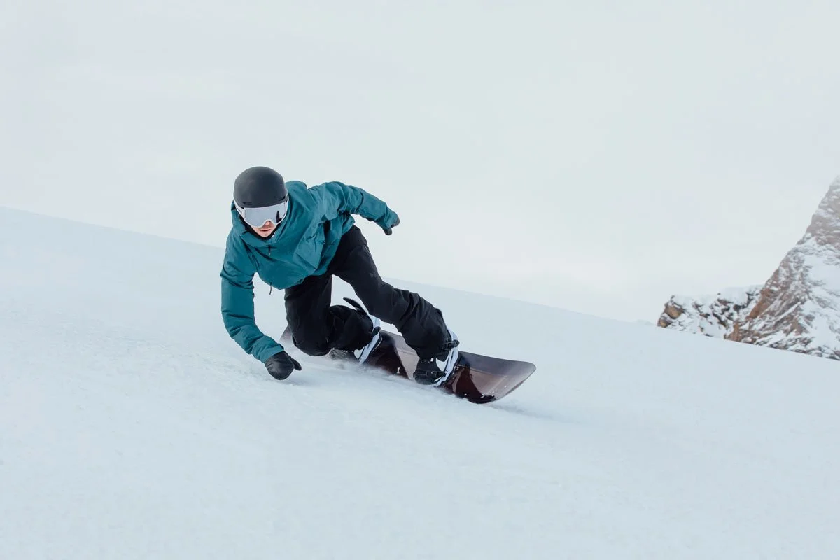 Skier in blue jacket and black pants wearing a helmet and goggles, skiing downhill on snow with mountain rocks in background.
